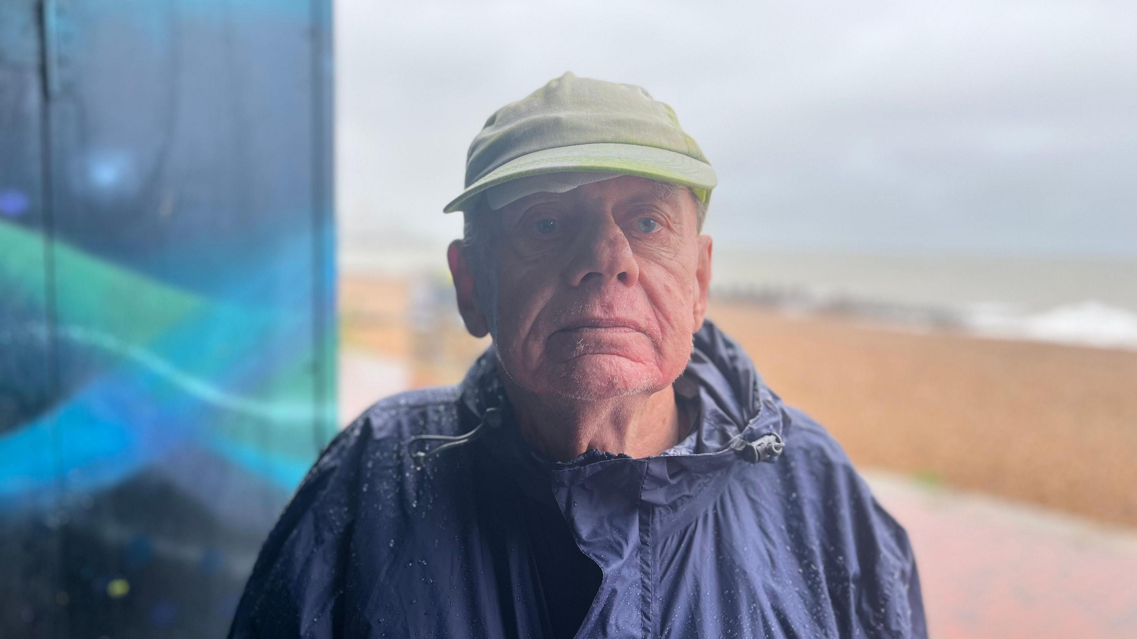 Alan Shiel is an elderly white man. He is wearing a navy rain coat, and a light-coloured hat.