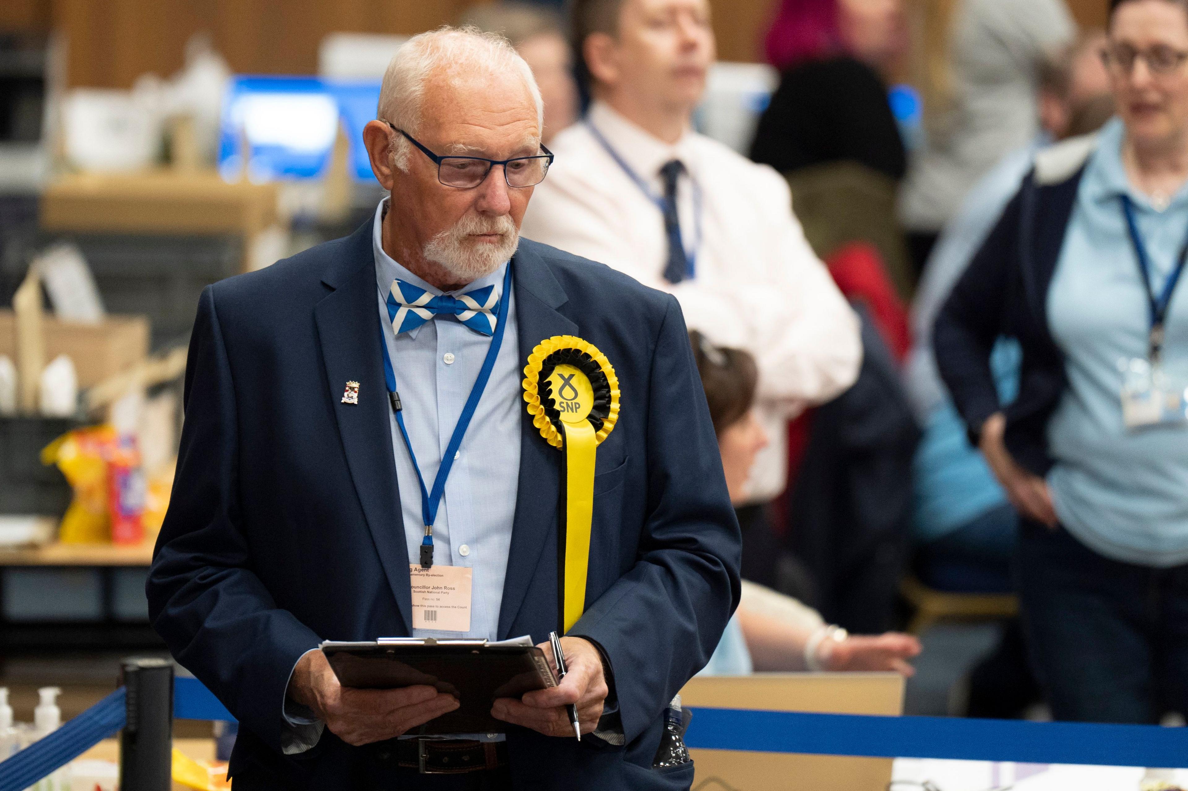 A serious looking SNP counter in Saltire bow-tie and blue suit stands with a clipboard watching the vote tally in a hall full of people.