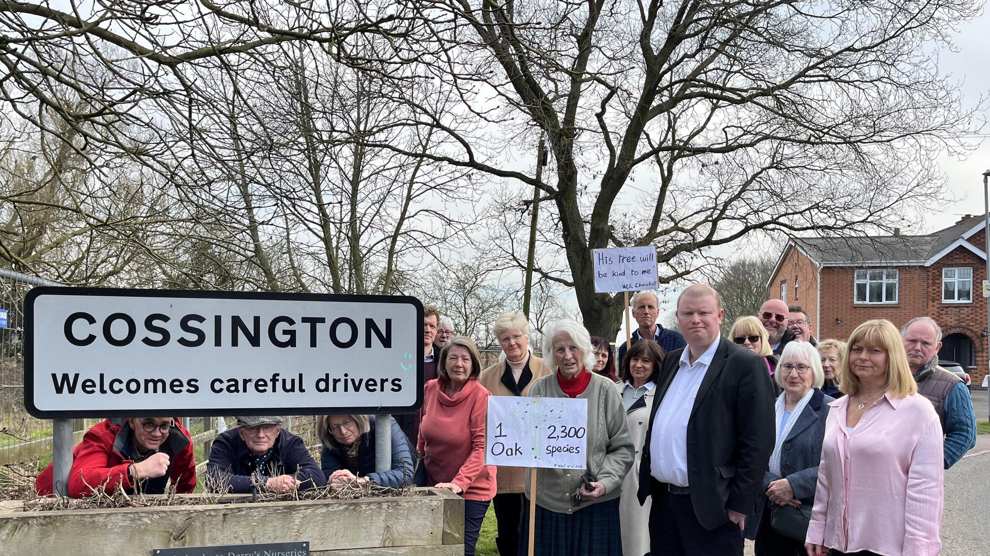 About 20 people from the village stand in front of the tree and a Cossington Road sign 