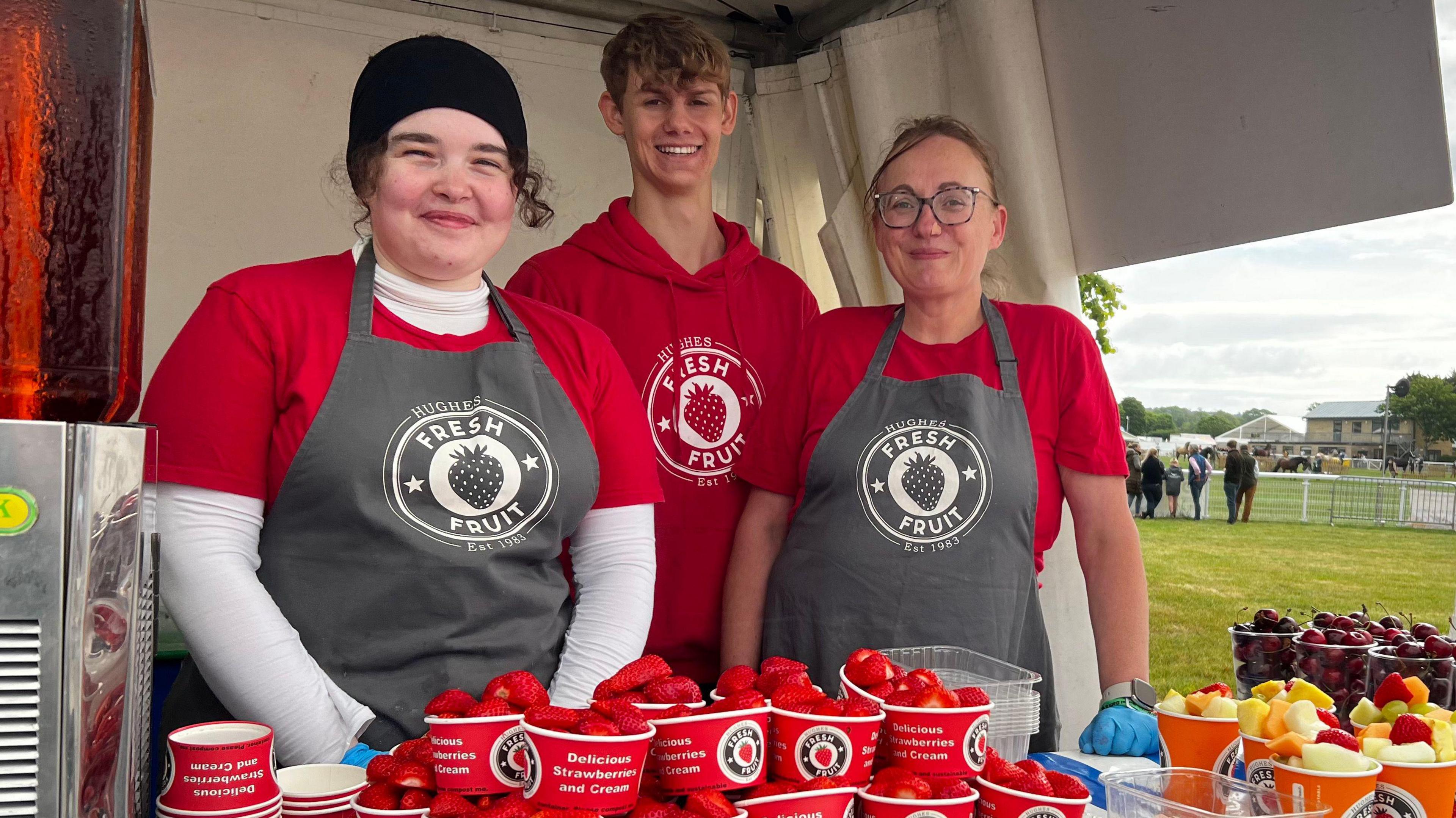 Three people wearing red behind a stall counter at the Bath and West Show smiling at the camera with small boxes of strawberries and other fruits sitting in front of them. On their aprons, there is a circular logo, with the words "fresh fruit" written above and below a picture of a strawberry.