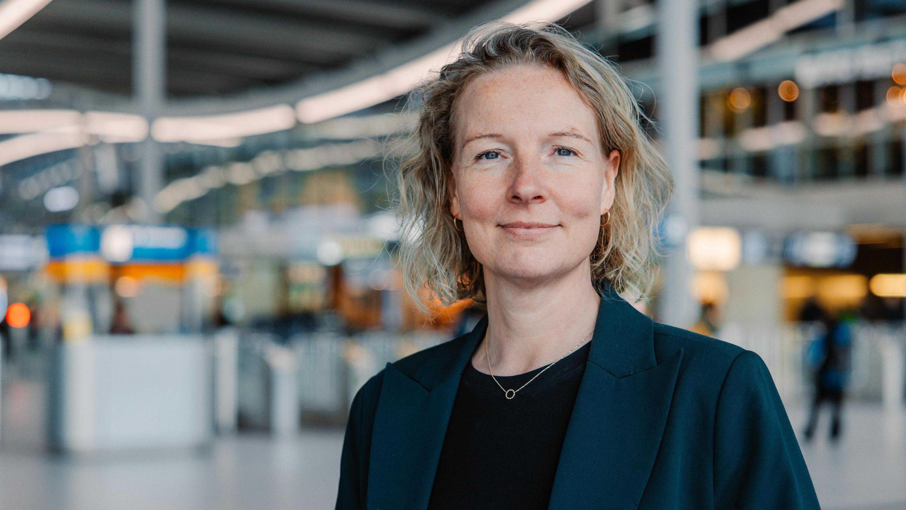 Bertien van Baak pictured in railway station smiling. She wears a black top and a navy blazer and has curly blonde hair