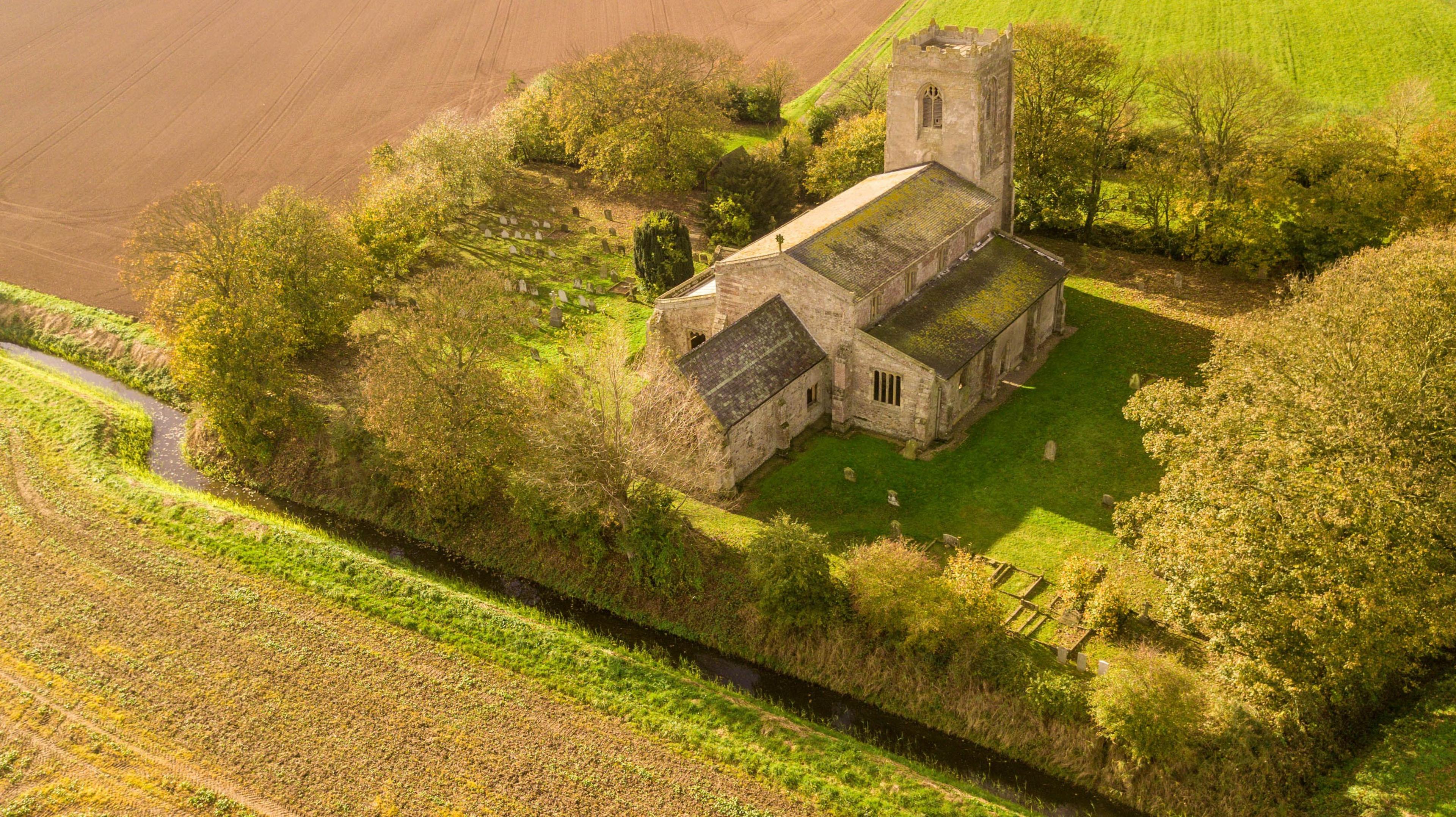 A drone view of a medieval church with tower in neat grounds, with a grassed burial ground surrounded by trees and fields and a narrow drain running past in the foreground.