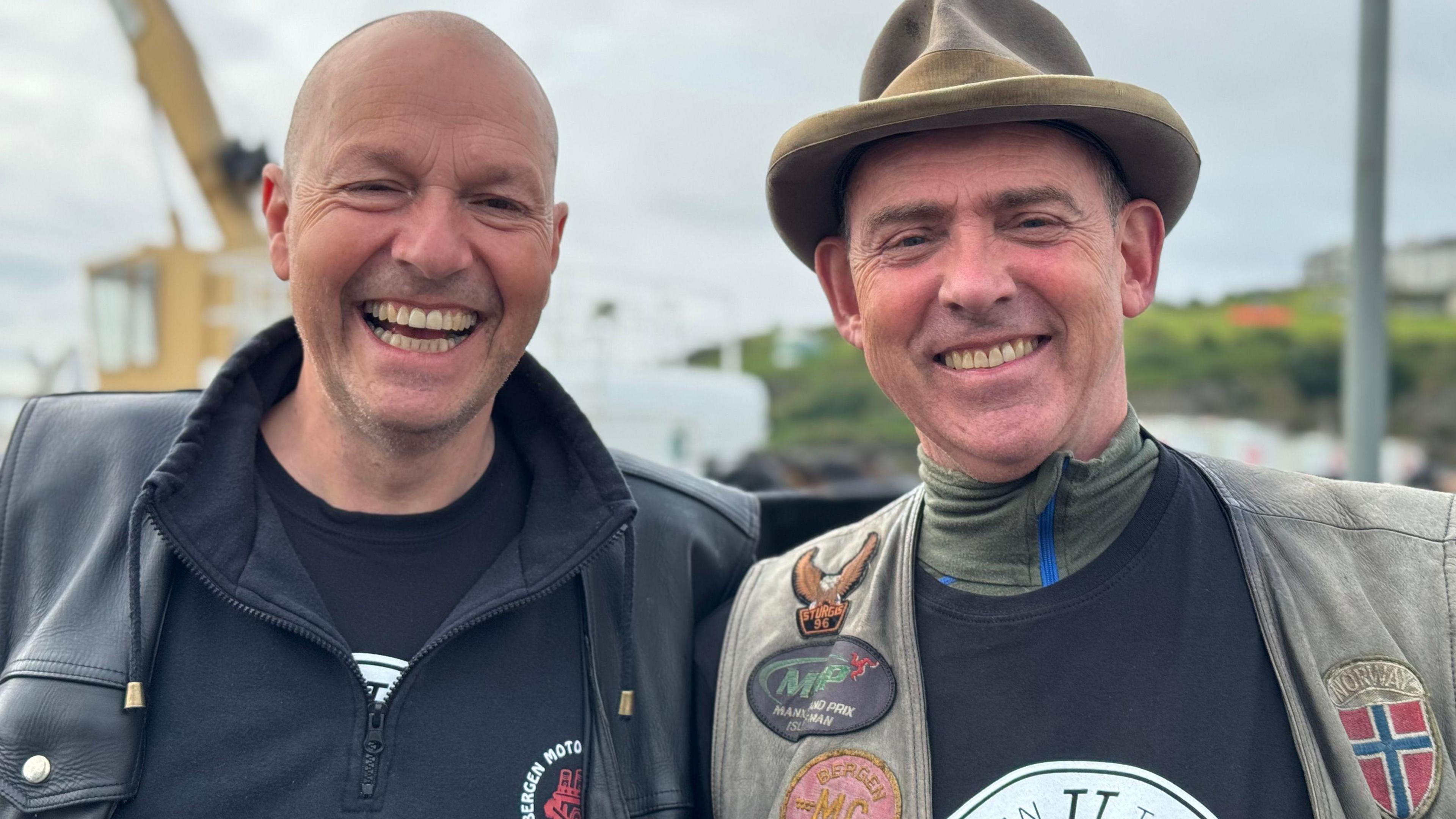 Ole Skaar Erling and Thomas Pettersen standing next to each other with smiling. Ole is bald and is wearing a black leather coat and black hooded jumper. Erling is in a black top and green waistcoat covered with badges and a camel-coloured hat.