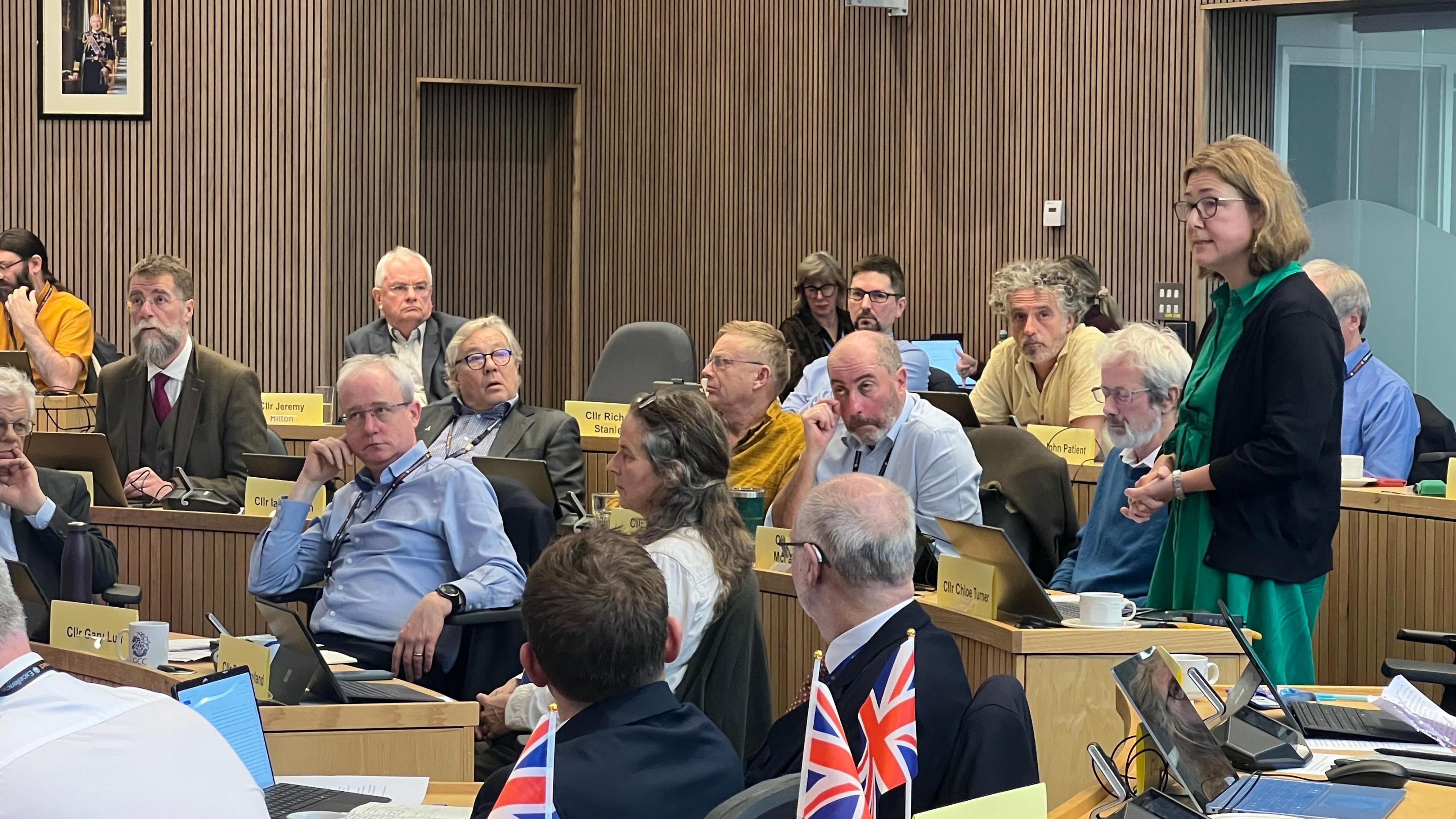 A large group of councils sit at desks in a circular council hall. Some have union jack flags on their desks. A woman in a green dress and black cardigan and shoulder-length hair is standing up addressing the room. 