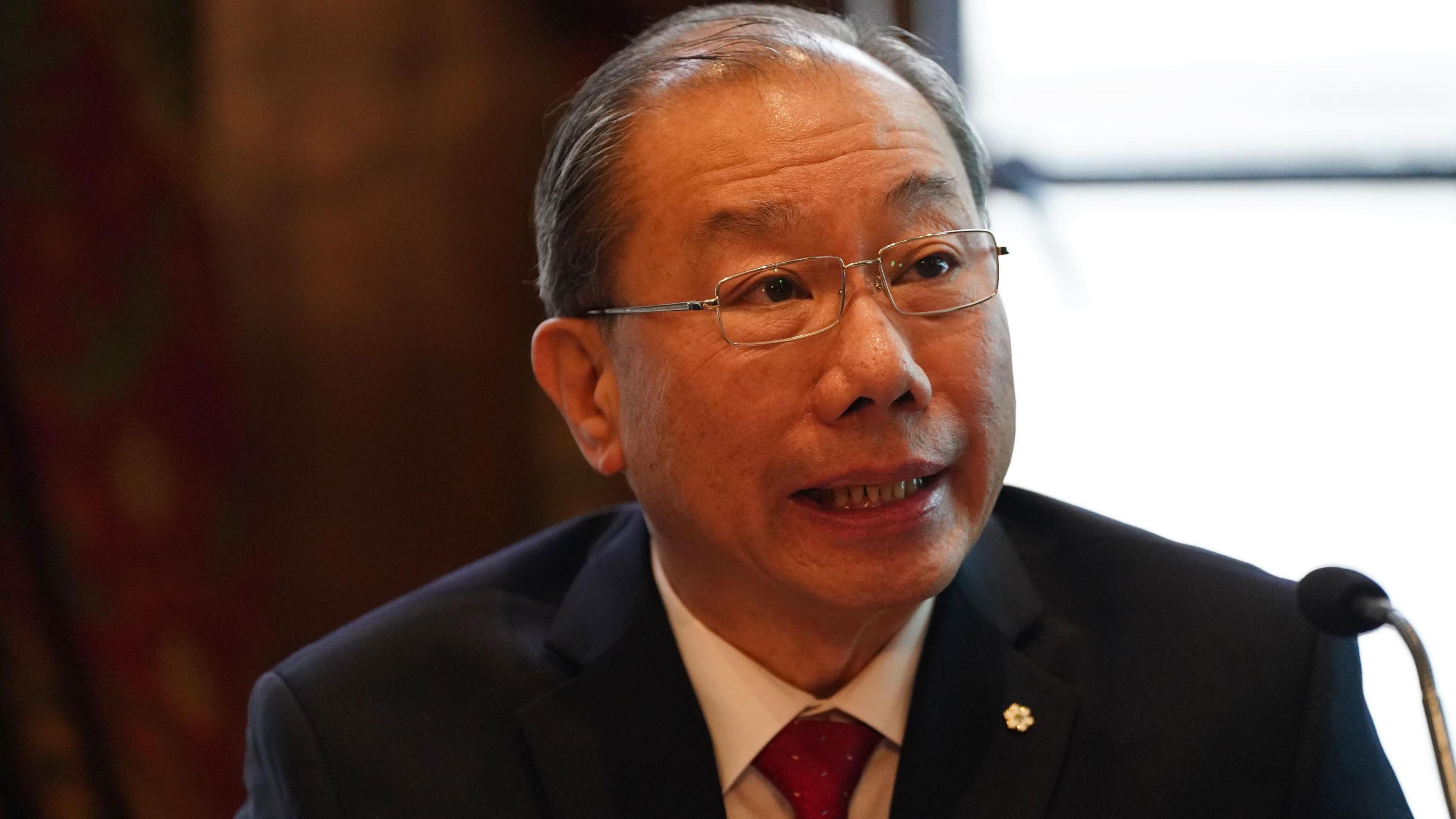 Dr Shoo Lee, who has thinning black hair and wears rectangular-framed spectacles, is wearing a navy-blue suit over a white shirt, speaks into a microphone.