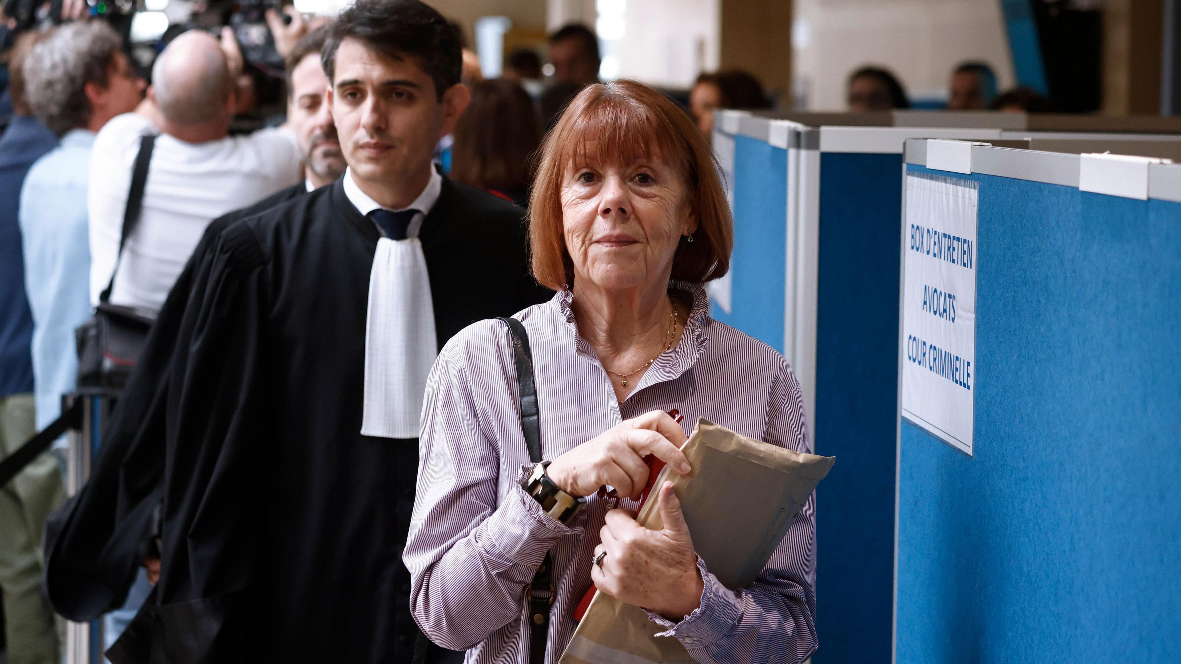 Gisèle Pelicot, a woman with a red bob and a purple blouse carrying a folder of documents, is walking through a room, followed by Stéphane Babonneau, a man with dark hair in a formal black gown