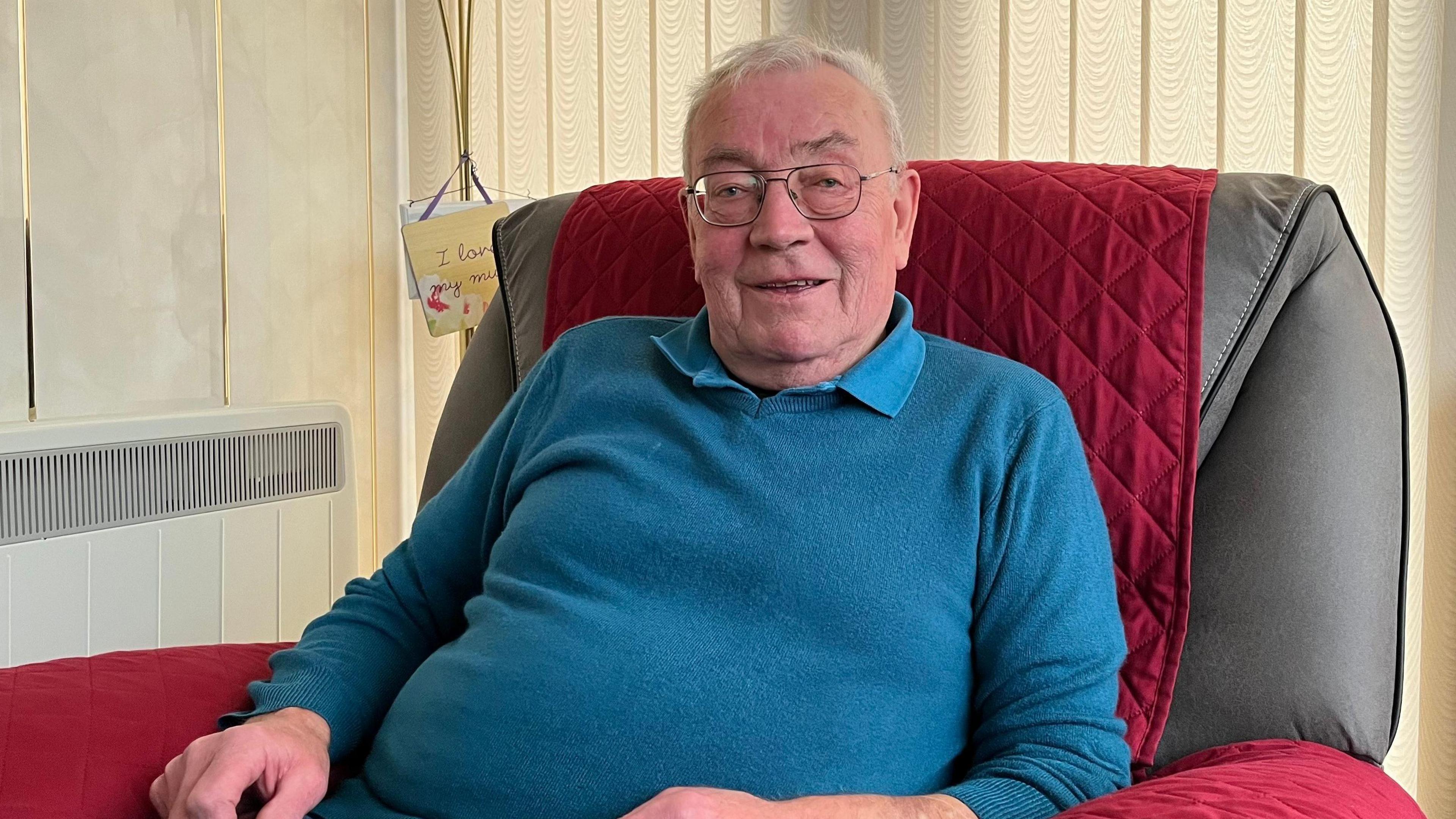 Murchadh MacIlleathain, air a bheil aillse a' phròstata agus e na shuidhe ann an cathair agus gàire air aodann. Tha geansaidh ghorm air agus plaide dhearg air a' chathair. 