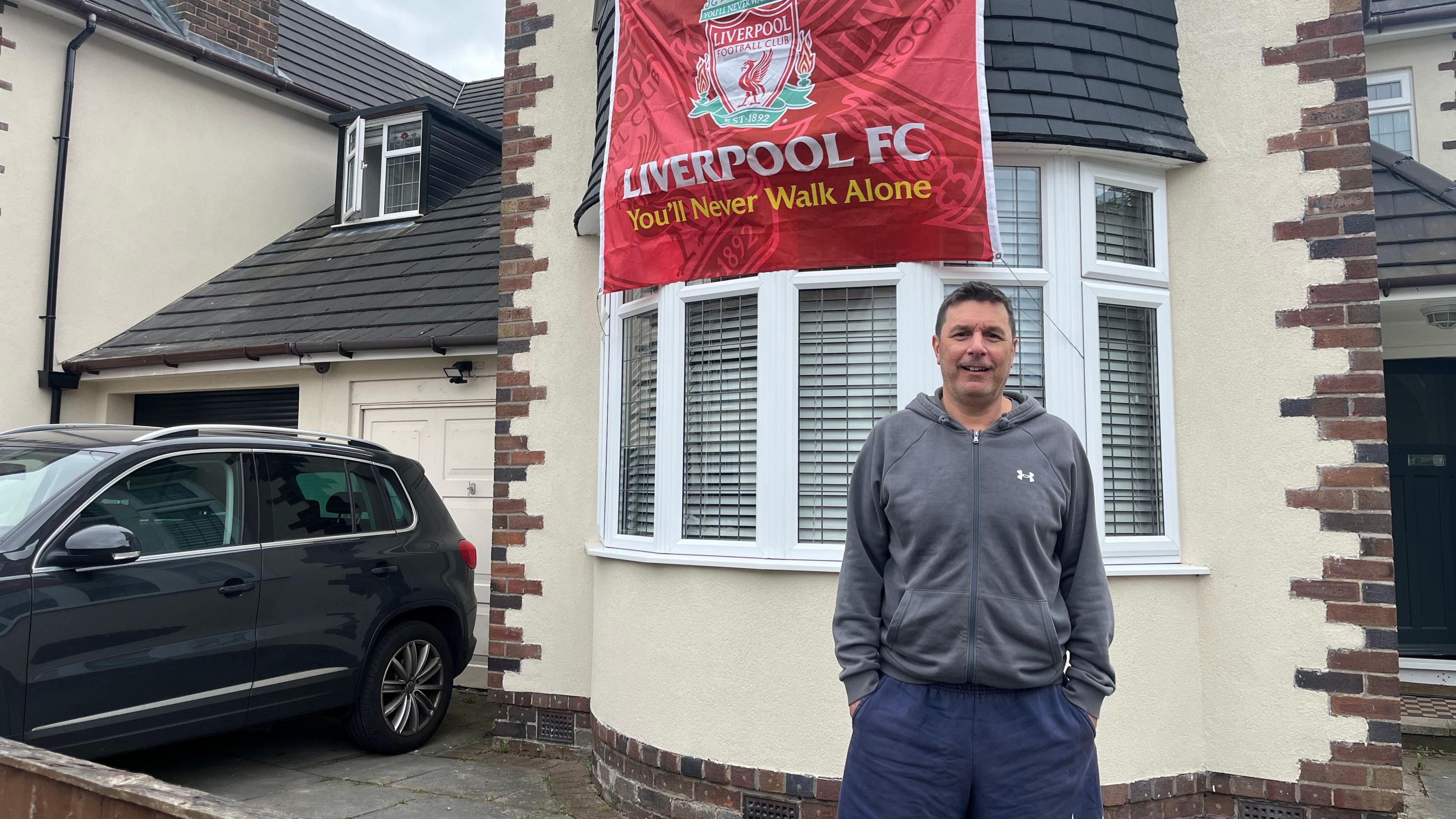 Chris de Asha, who has dark hair, a grey zip up hoodie and blue shorts, stands in front of a semi-detached house with a large red Liverpool FC banner hanging from an upstairs window.