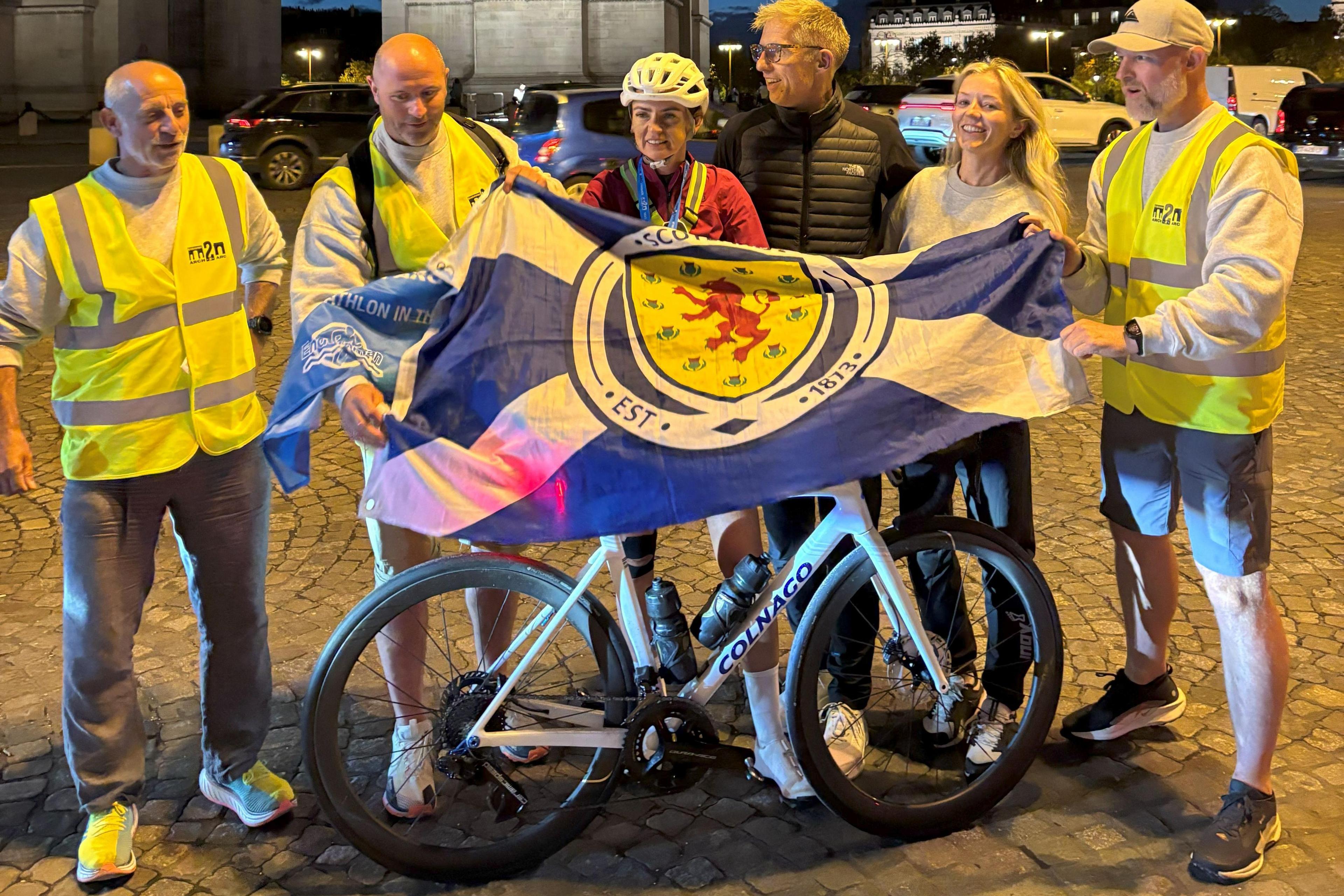 A smiling Lyndsey Blair waves a Scotland flag with her husband - who has grey hair, glasses and is wearing a black North Face jacket - niece - who has blonde hair and is wearing a grey sweater - who finish line in Paris. She is wearing black cycle shorts, a red long-sleeved cycle top and a yellow helmet. She has a medal round her neck