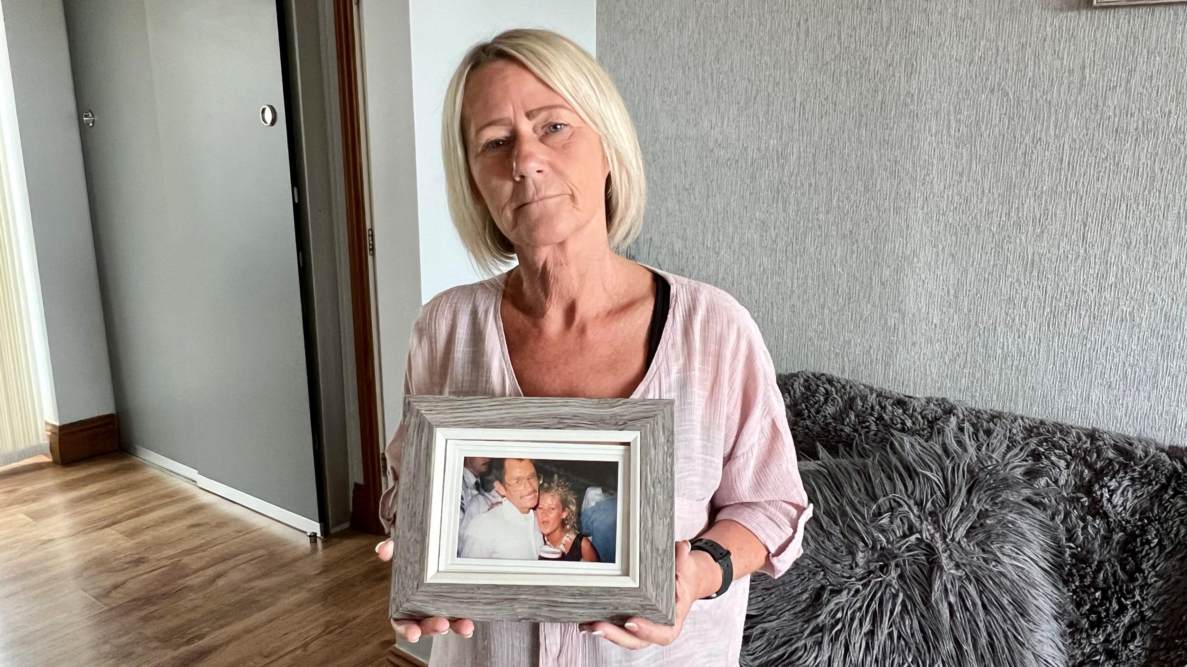 A woman in her fifties with blonde hair and a light pink top is holding a picture. It shows her as a younger woman hugging her dad. She is standing in her living room in front of a sofa.