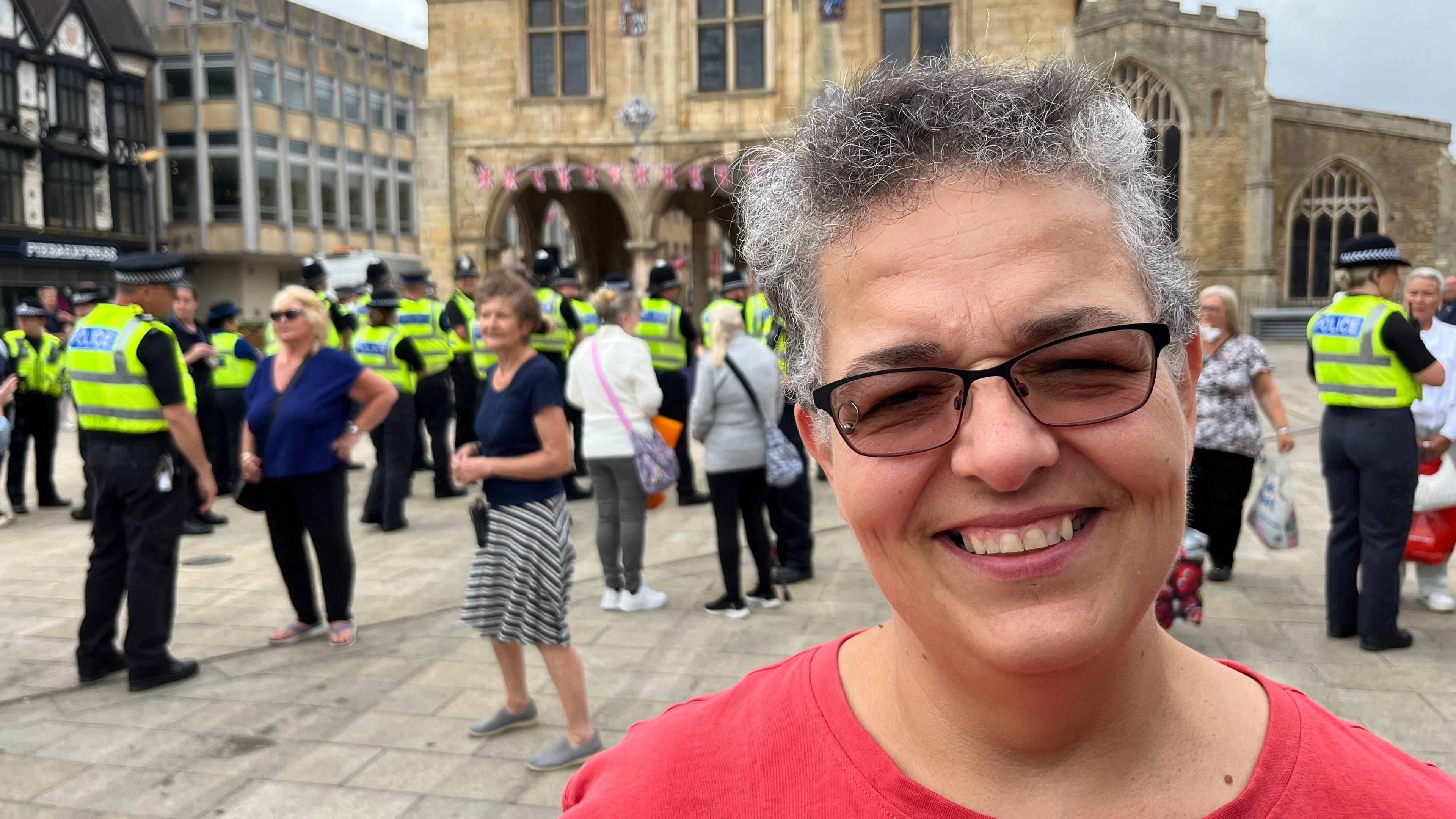 Woman wearing a red T-shirt stands in Peterborough square. She is smiling and looking at the camera, She has greying frizzy hair and wears glasses. Behind her are lots of police officers and members of the public. A church can be seen in the background and shops.