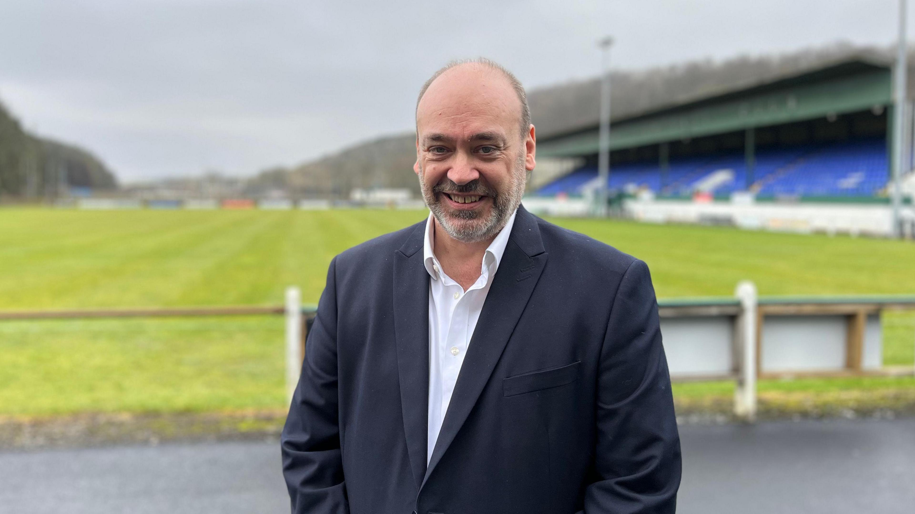 A bald-headed man with a grey beard, who's wearing a black jacket and white shirt, is smiling at the camera. A rugby pitch and stand are in the background