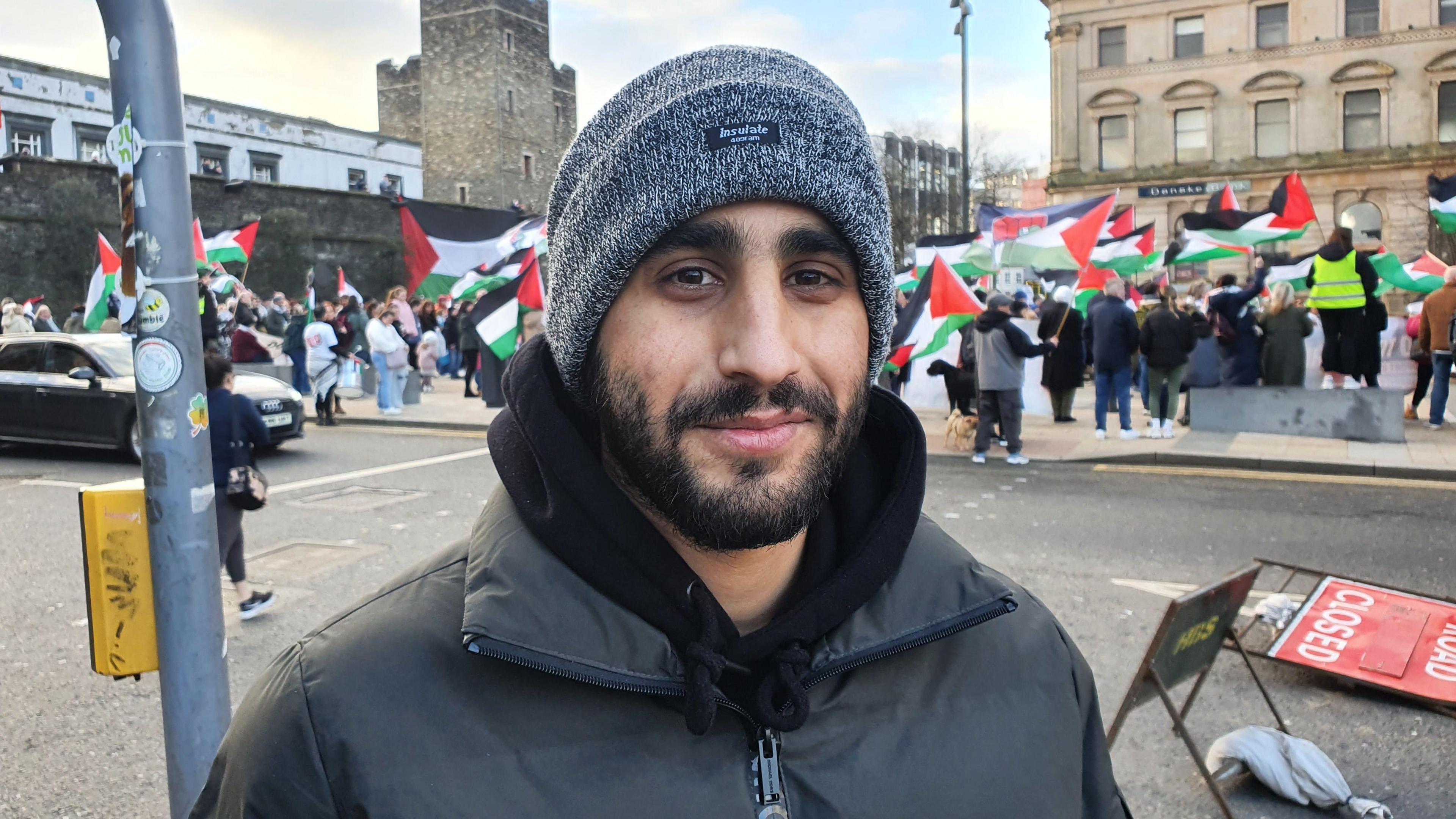 Mohyeddin Obeid smiles as he stands in front of a group of people. The group are holding Palestine flags. Obeid has on a grey woollen hat and black jacket with a black hoodie underneath. 