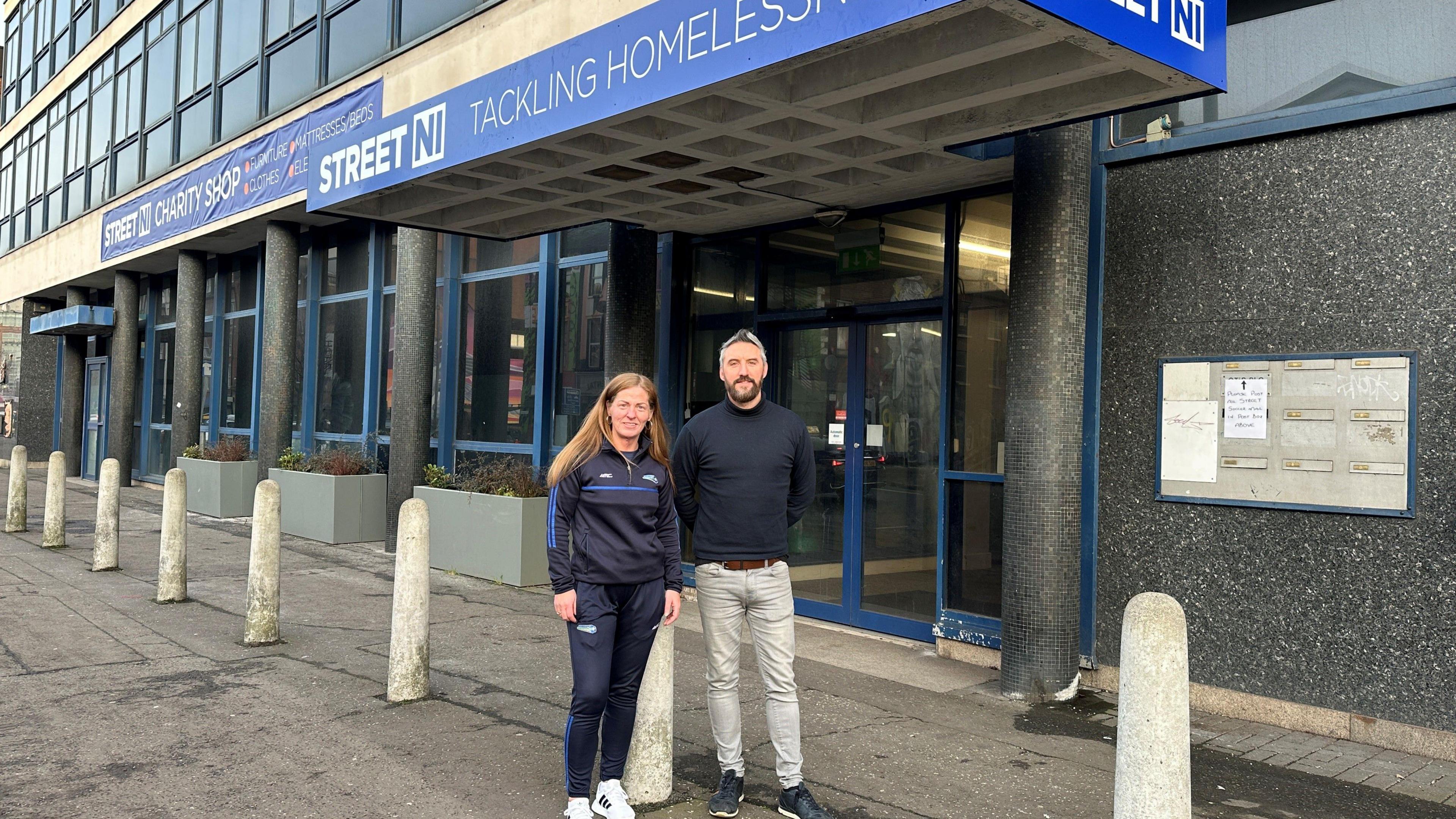 Louise, left, is standing in front of the new hub. She is wearing a navy football tracksuit. Justin, right, is wearing light coloured jeans and a navy turtle neck top. The new signing above the door is blue with white writing that says Street NI Tackling Homelessness.