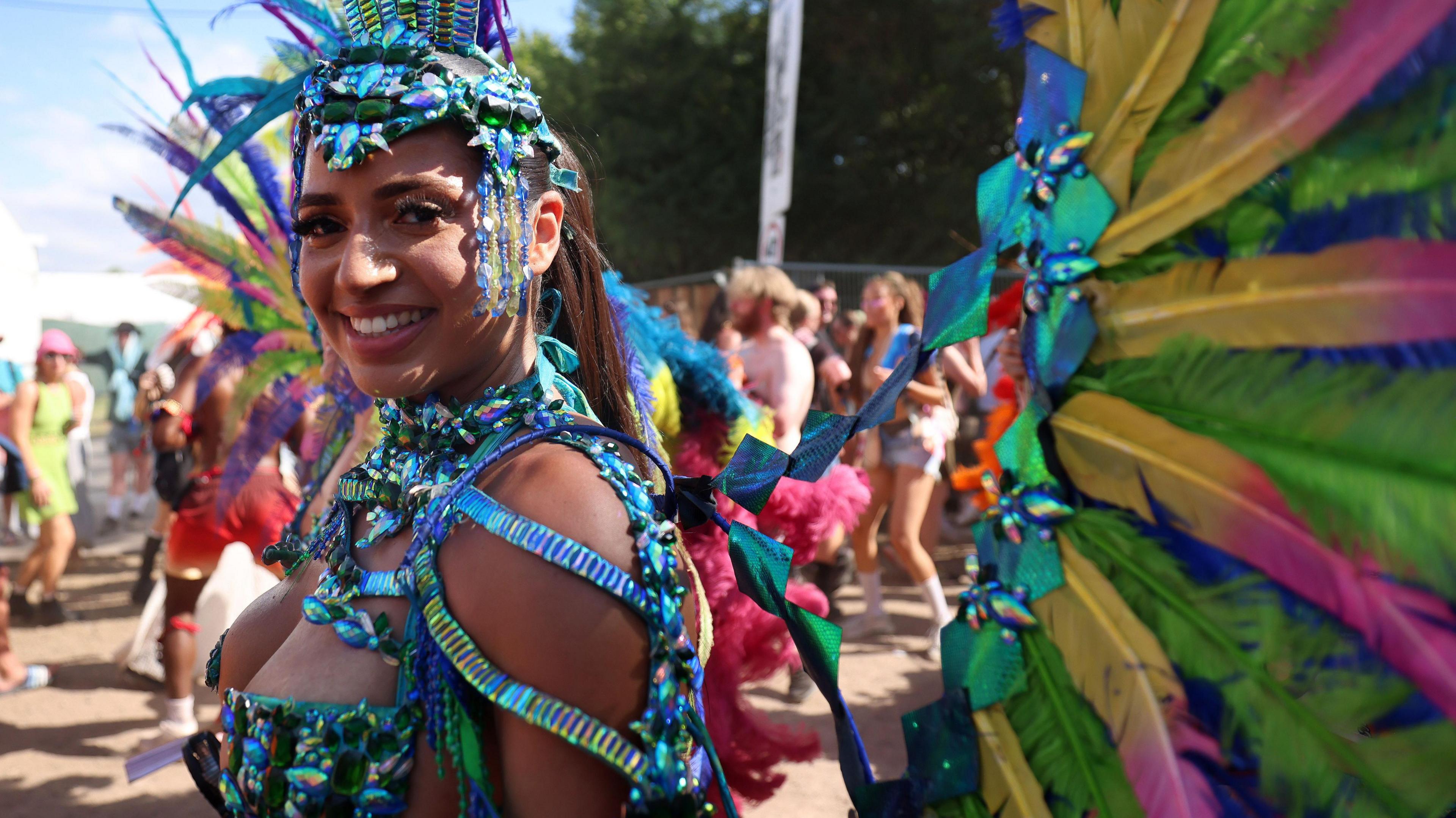 A woman is wearing a carnival costume, which is blue and glittery. She is also wearing wings made of colourful feathers. 