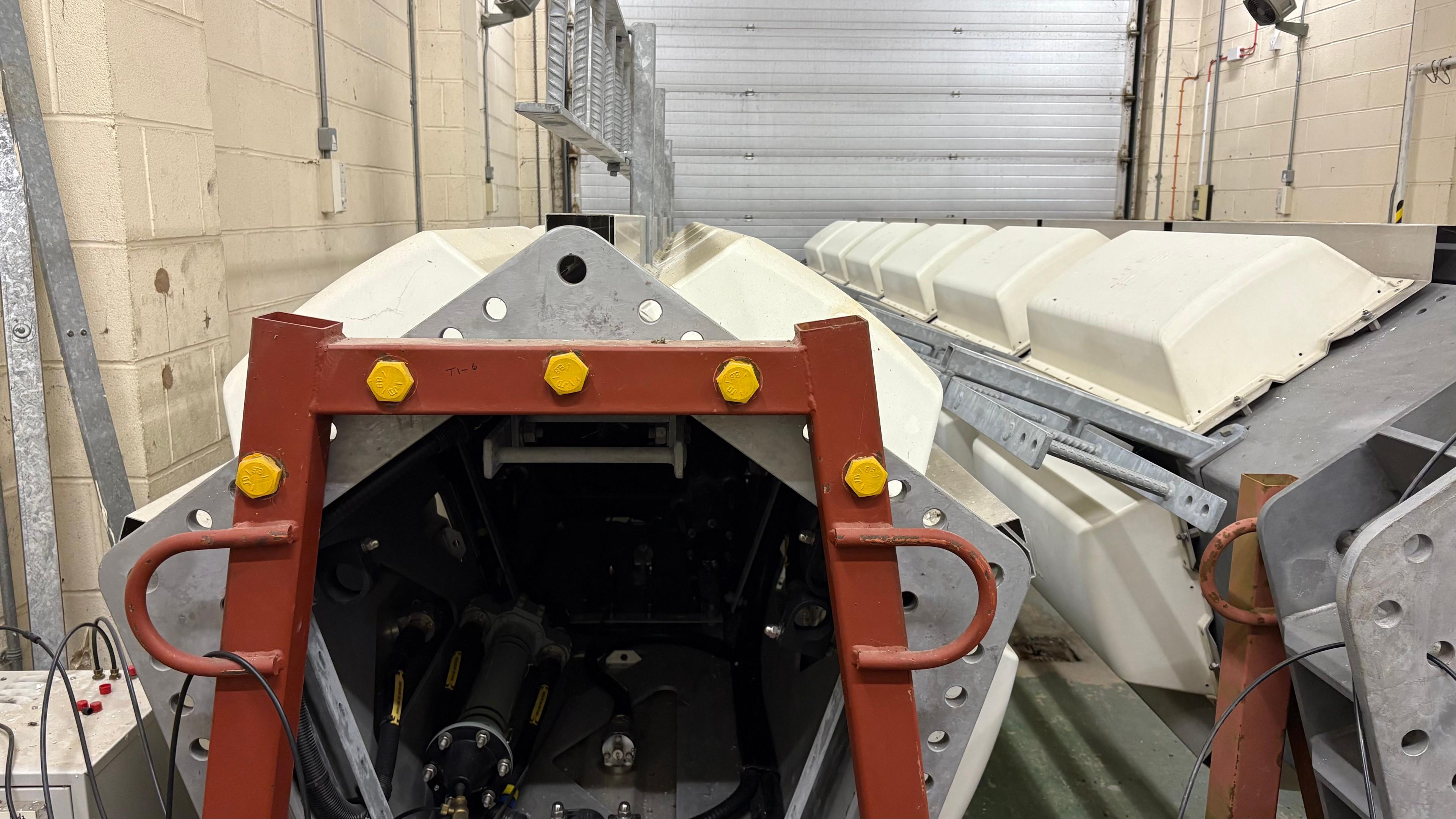 Two pentagonal mast sections lying on a warehouse floor - the hollow side of one can be seen, surrounded by a red frame with gold-coloured bolts. Both sections have a line of white panels along the sides.