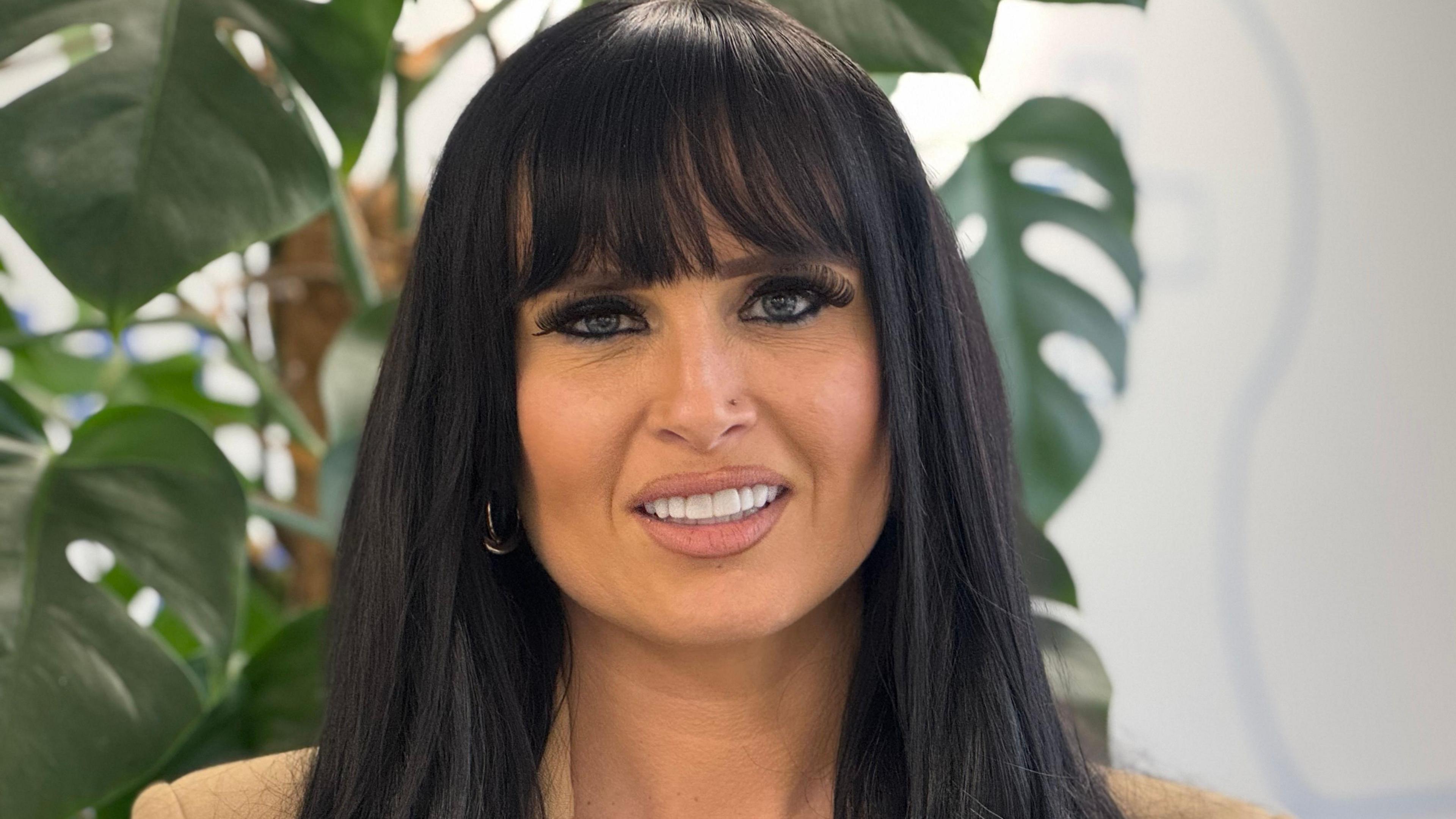 A woman with long black hair past her face looks into the camera with her mouth open, showing her teeth. Behind her is a plant with large, green leaves.