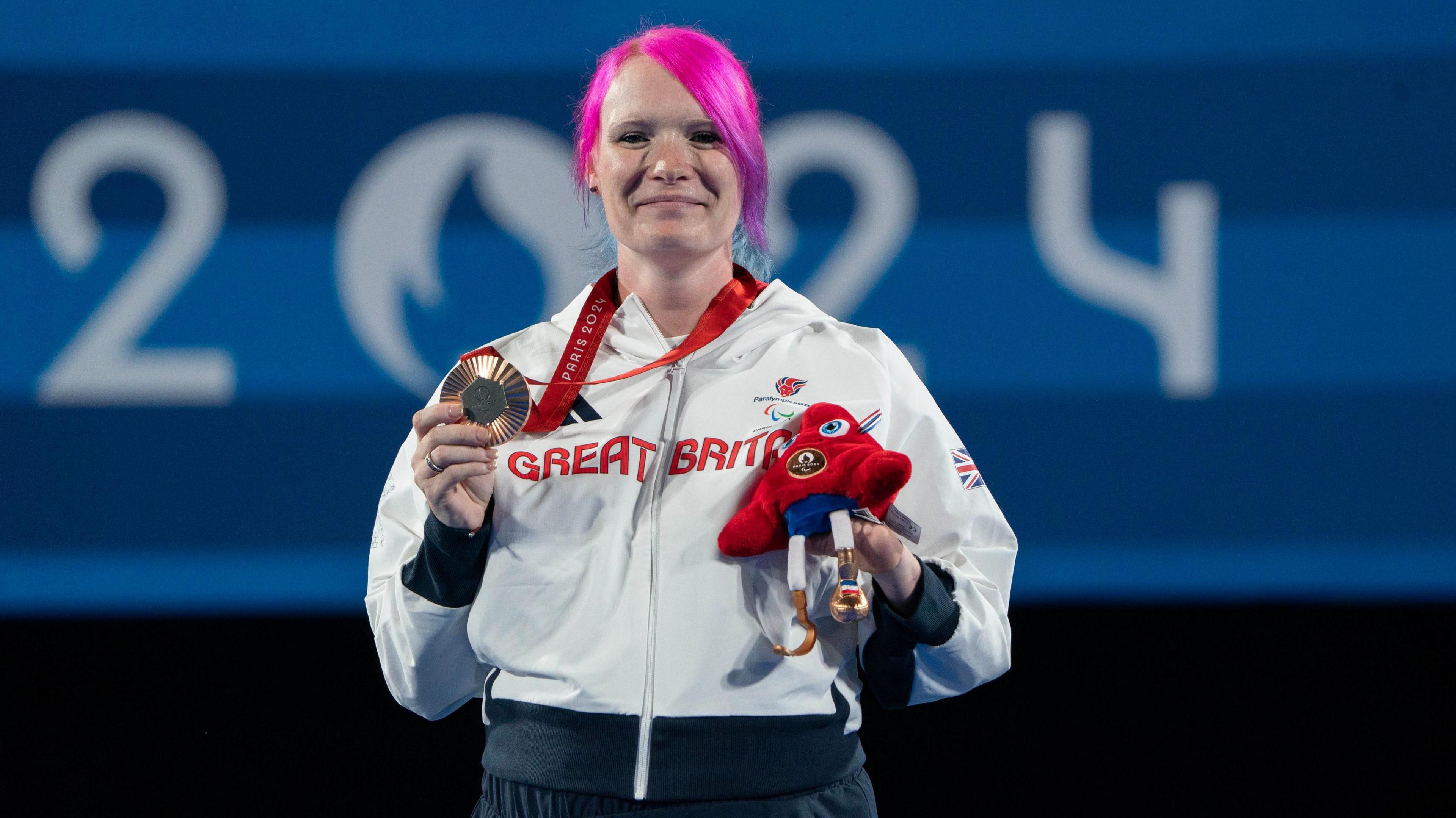 Great Britain's Jodie Grinham on the podium with the bronze medal after the Women's Individual Compound Open Bronze Medal event at Invalides during day three of the Paris 2024 Summer Paralympic Games. She has pink hair and is holding her medal in her right hand. 