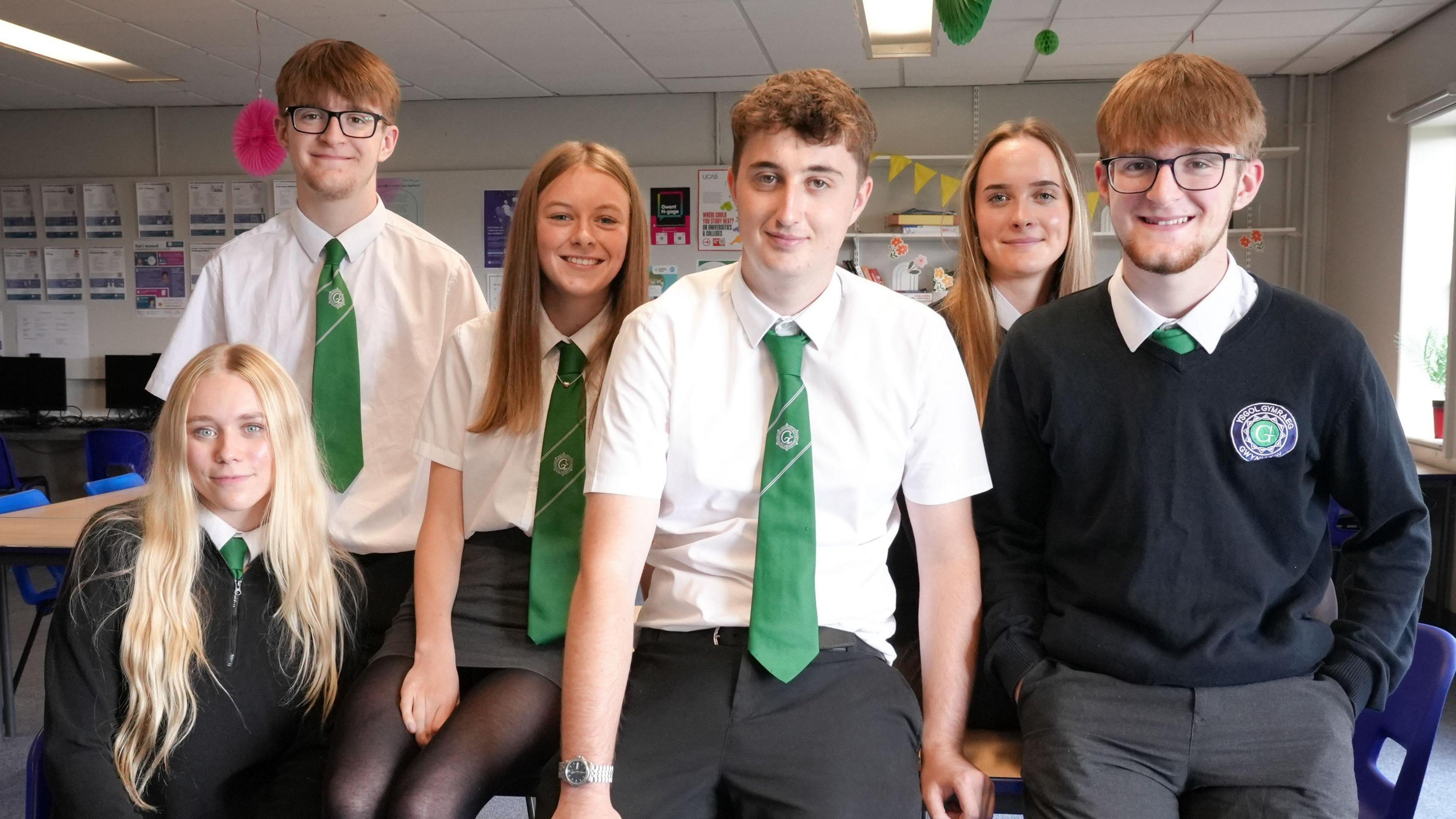 Tri bachgen a tair merch mewn gwisg ysgol gyda chrysau gwyn a teis gwyrdd yn eistedd ar y ddesg ac ar gadair mewn ystafell ddosbarth