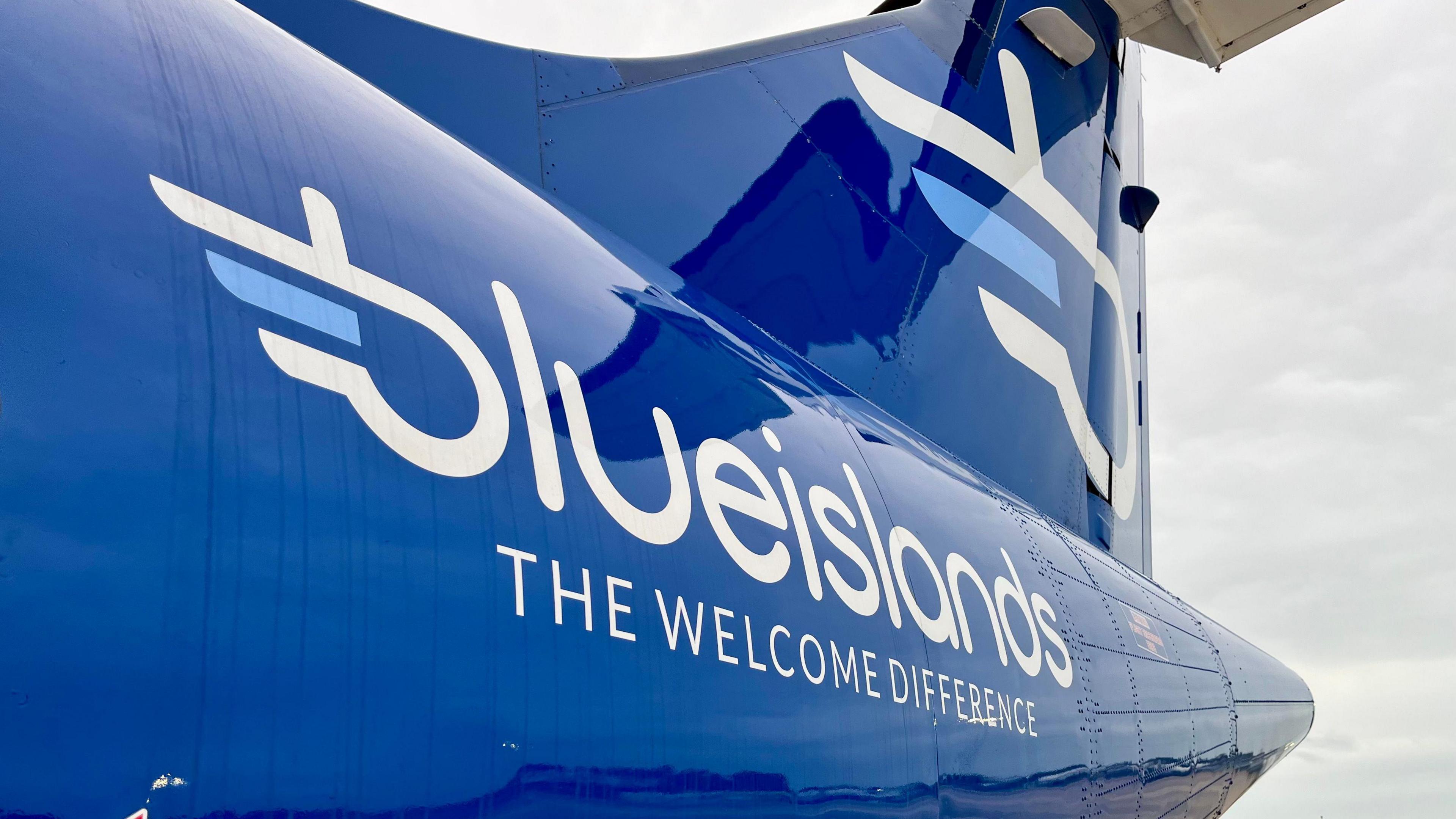 The tail of a plane, in royal blue. It says 'Blue Islands,The Welcome Difference' in white writing. Above the writing, there is a white and light blue logo. To the right of the image, the sky is grey.