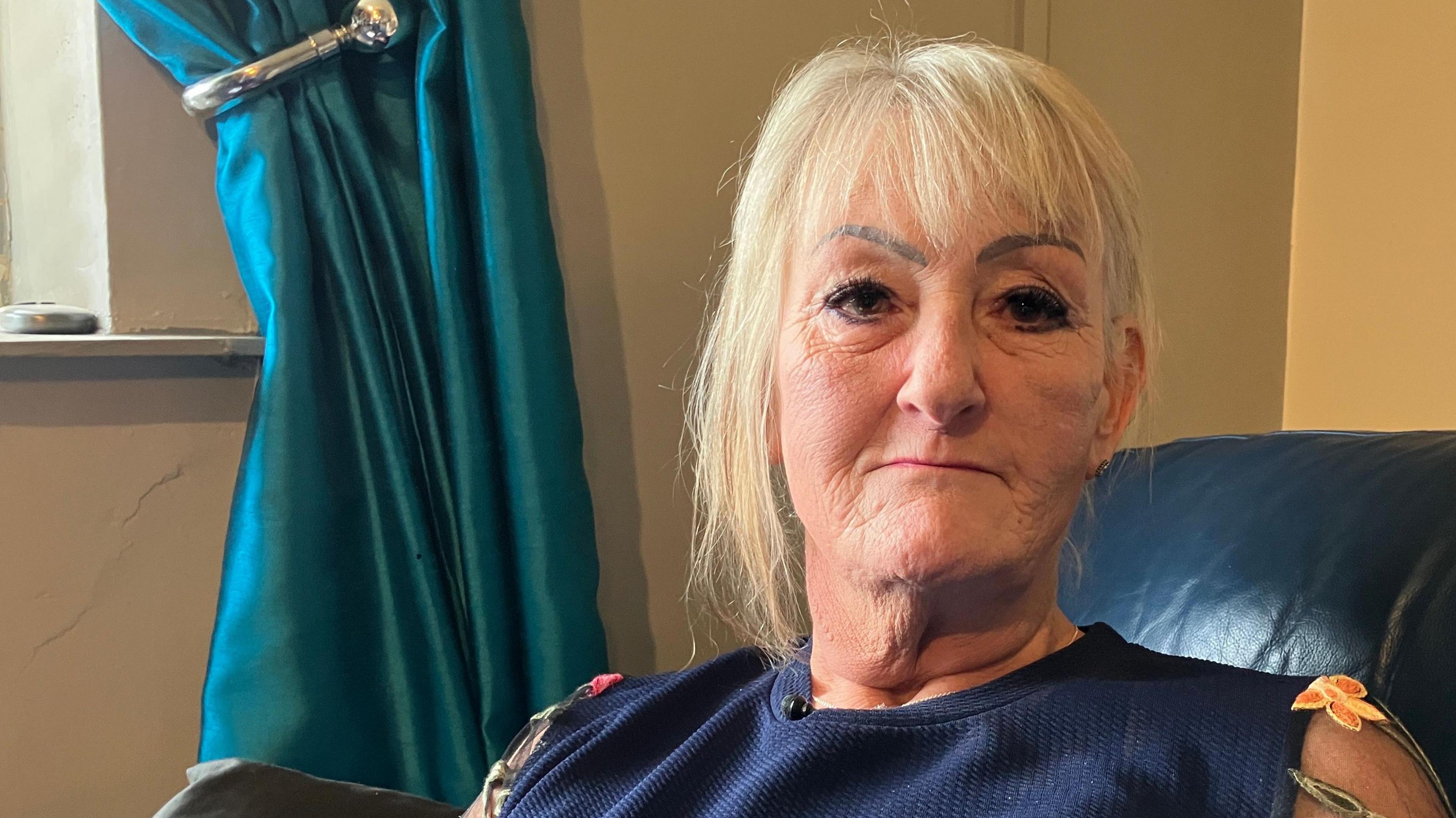 Maggie, who has blonde hair with a fringe, wearing a blue top with flowers on the arms. She is sitting on a blue leather sofa with blue curtains in the background.