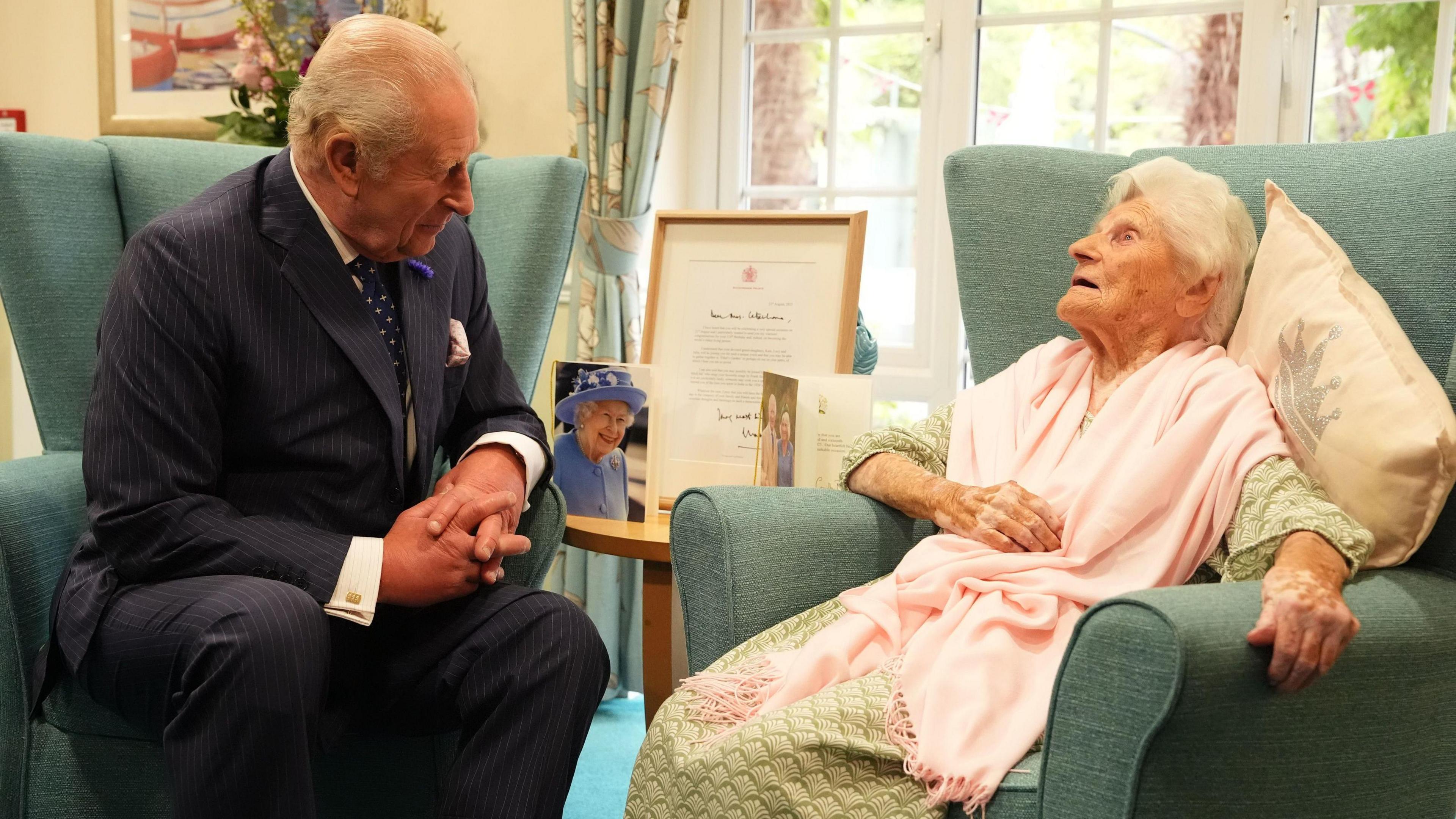 King Charles III meets the world's oldest person, 116-year-old Ethel Caterham, at Hallmark Lakeview Care Home in Lightwater, Surrey. Both are seen sitting in armchairs talking. 