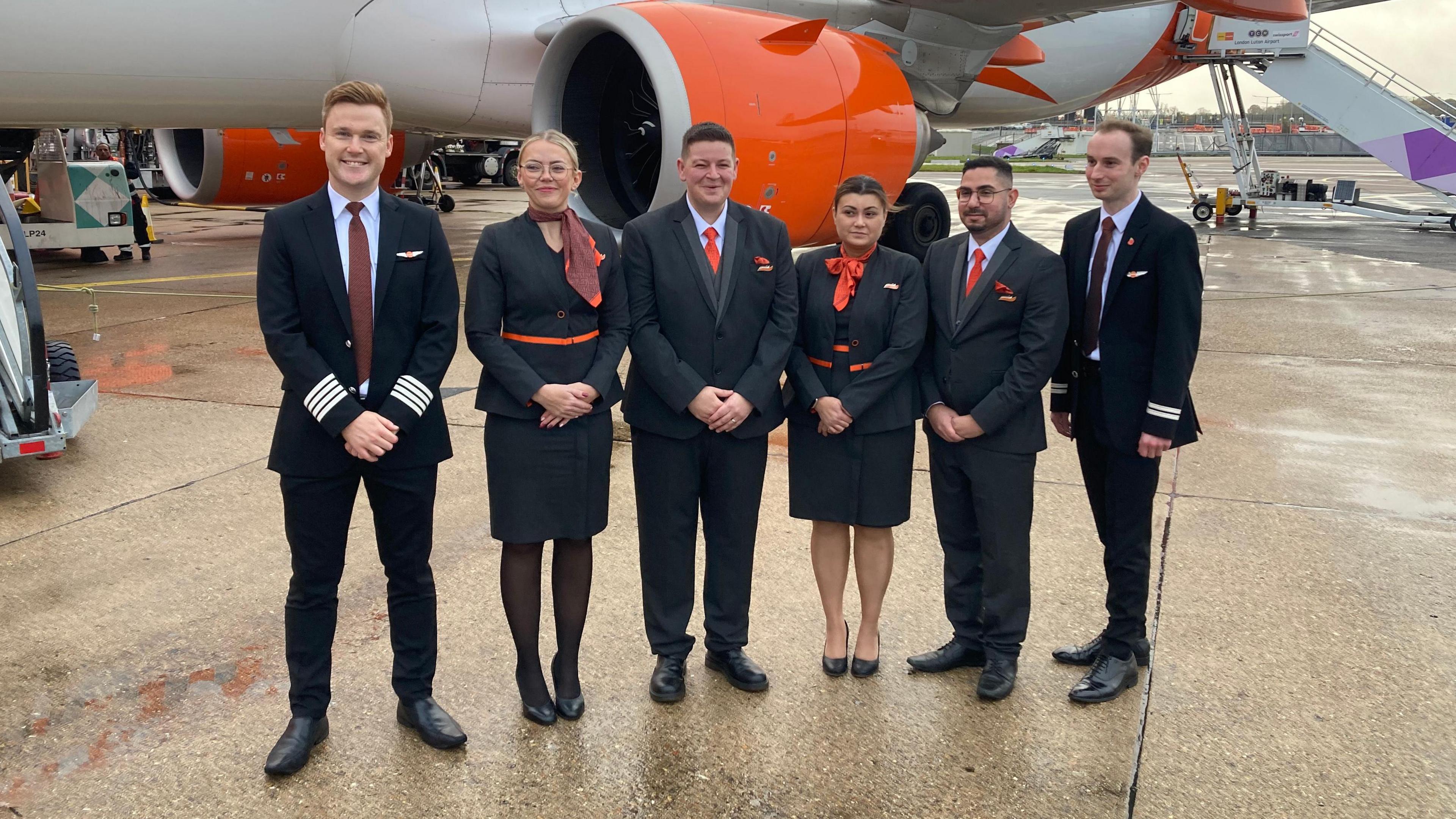 A group of airline crew are gathered outside in front of a white and orange aeroplane