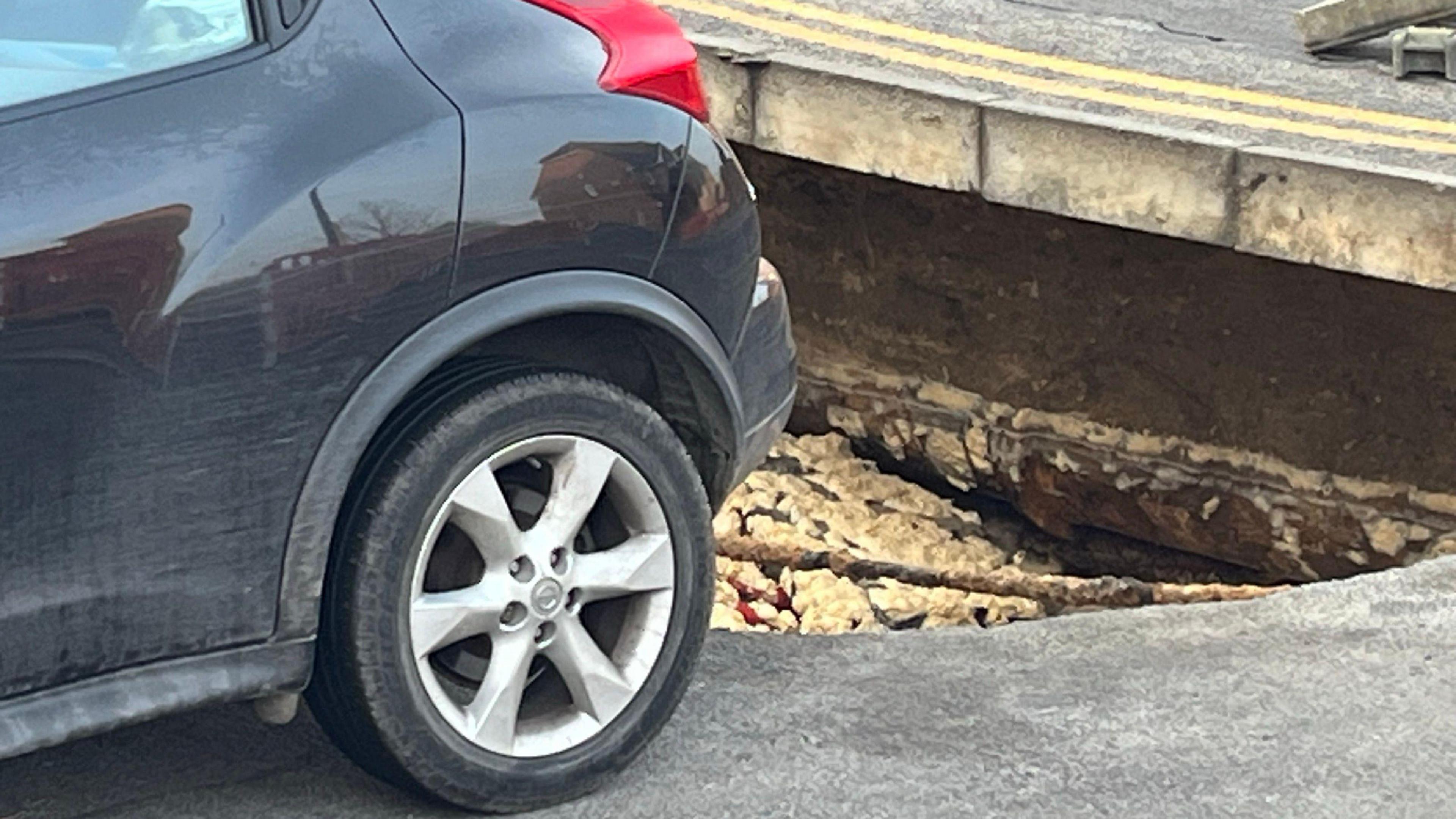 A dark blue car parked in a drive with a crack caused by a sinkhole reaching up to the rear wheel.