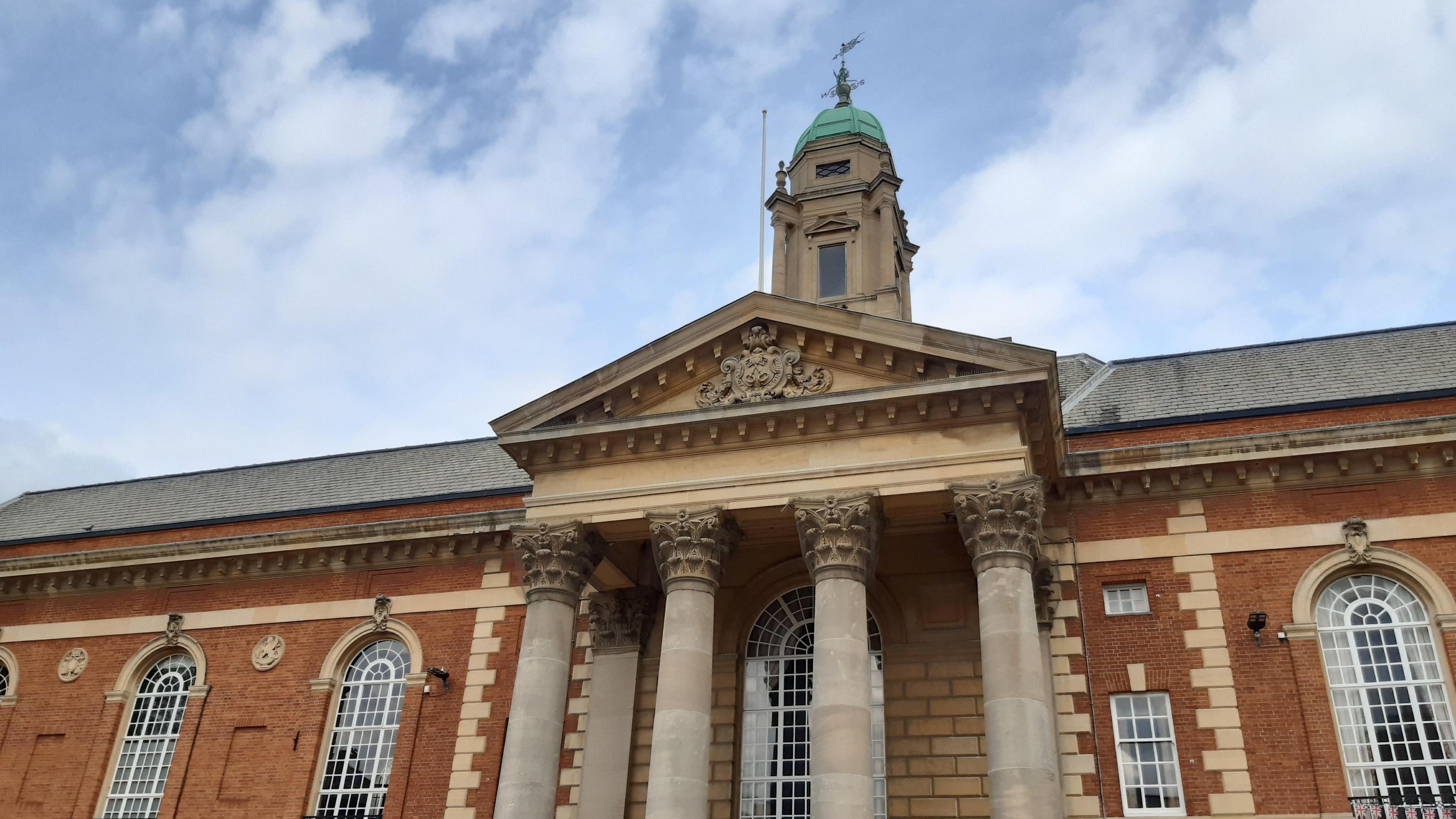 A close up of Peterborough Town Hall. It is a brown brick building with 4 pillars in front. A number of arched windows are also on the building.