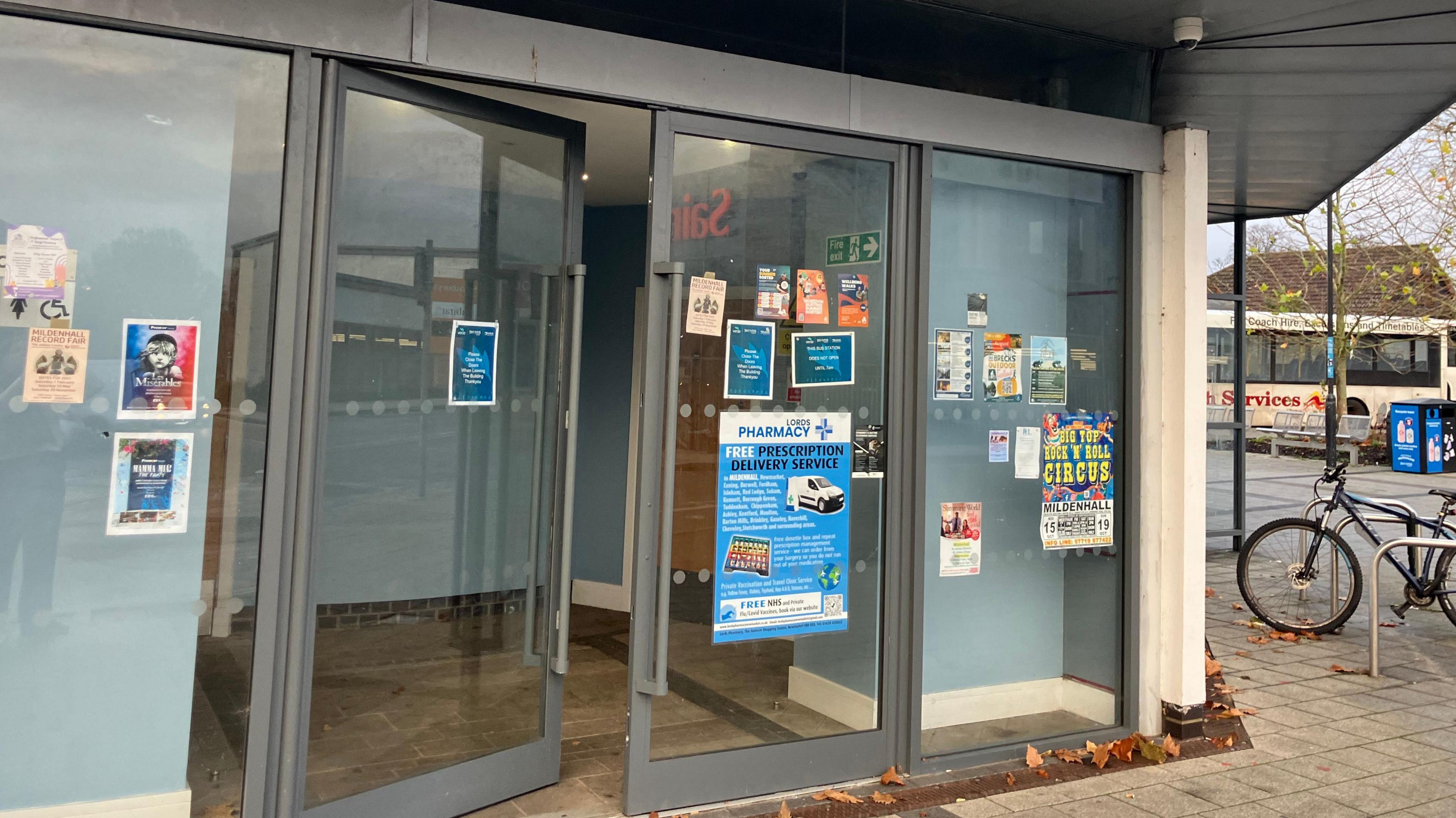 A close-up of floor-to-roof metal-framed glass doors, with a number of posters and flyers stuck to them. Above is a jutting-out roof ledge. To the right, a bike tied to a bike rack is just in shot, and beyond it there is a white single decker bus
in the distance.