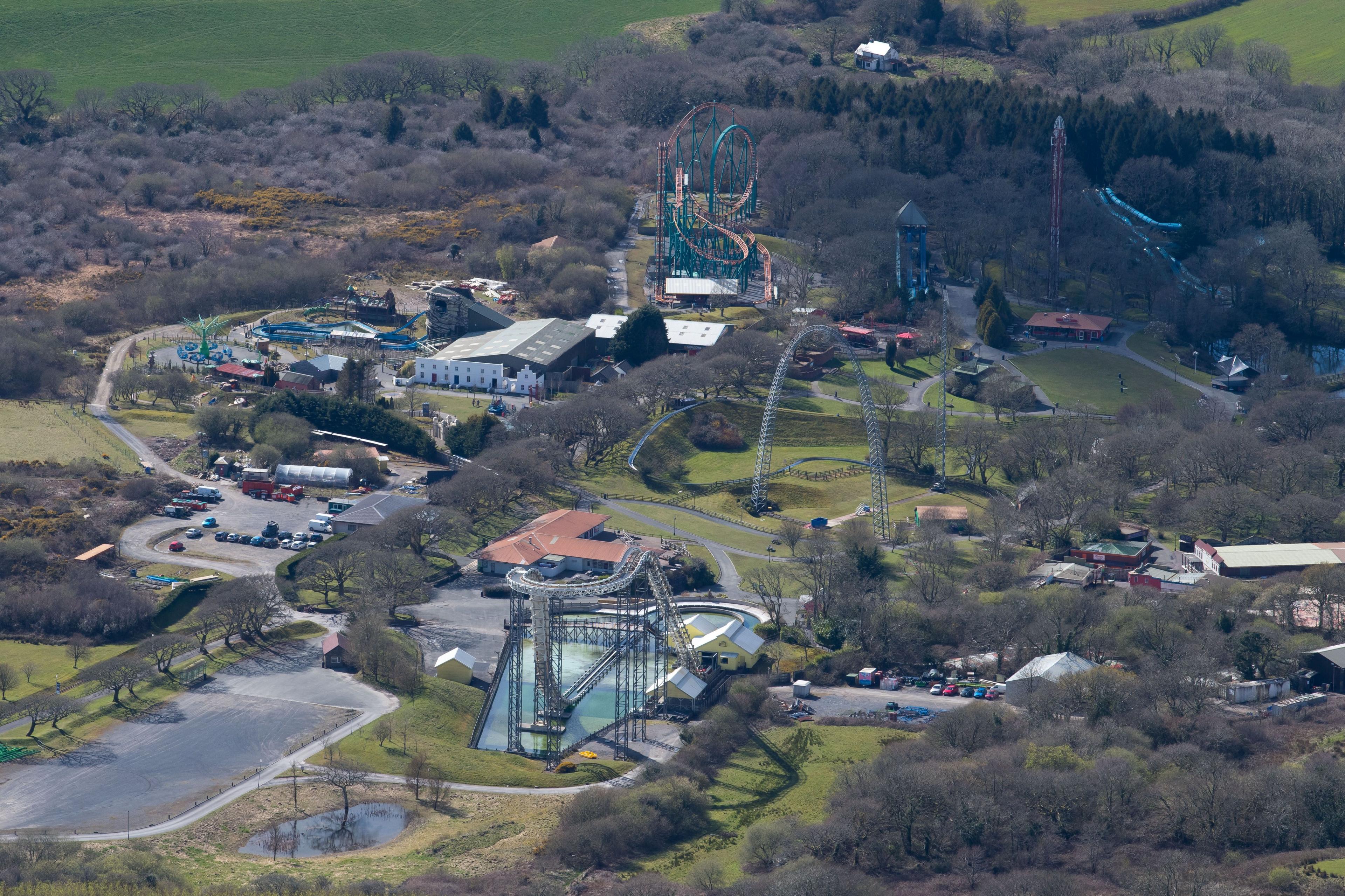 An aerial view of Oakwood theme park. A number of large rollercoasters can be seen spread across the park, with grass and trees in between the attractions. A large white building can be seen at the back of the park. 