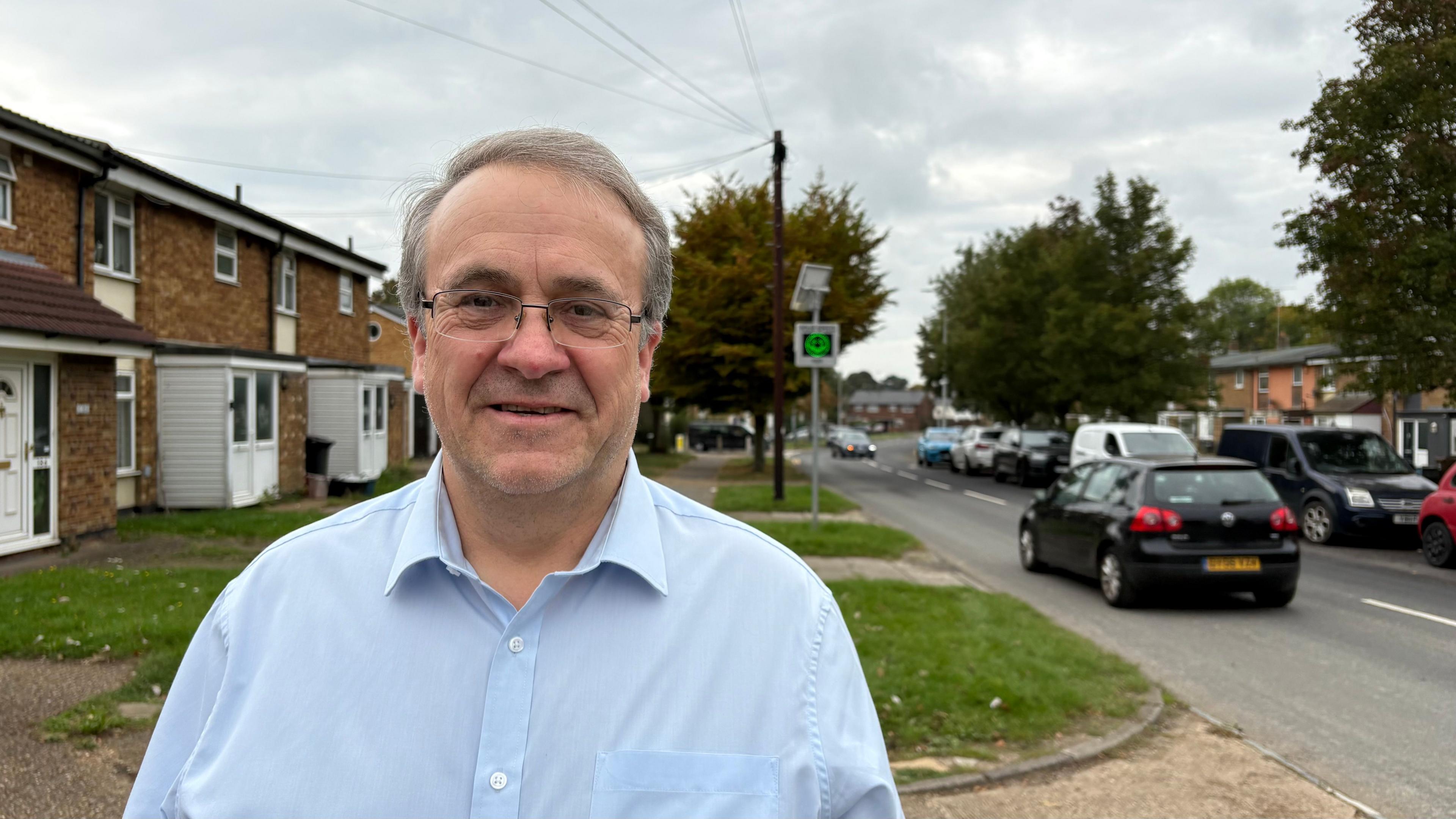 Paul Zukowskyj with light blue shirt, short white hair and glasses looking at the camera with two storey houses behind him, with grass at the front. There is a road to his left with cars parked alongside the pavement and a black car travelling away from the camera.