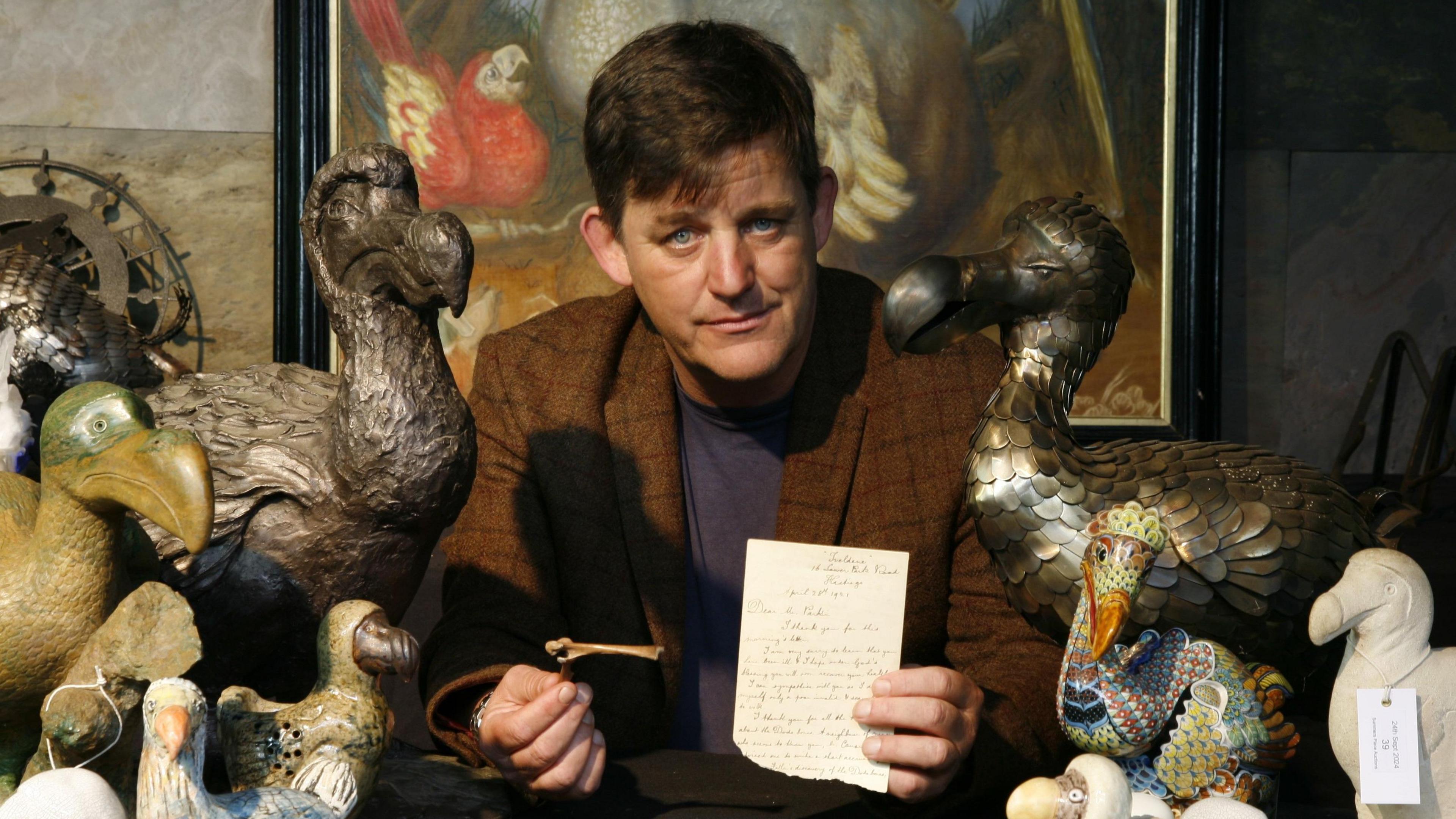 A man in a brown jacket is sat at a table surrounded by dodo sculptures. He is holding a fragment of bone and a letter.