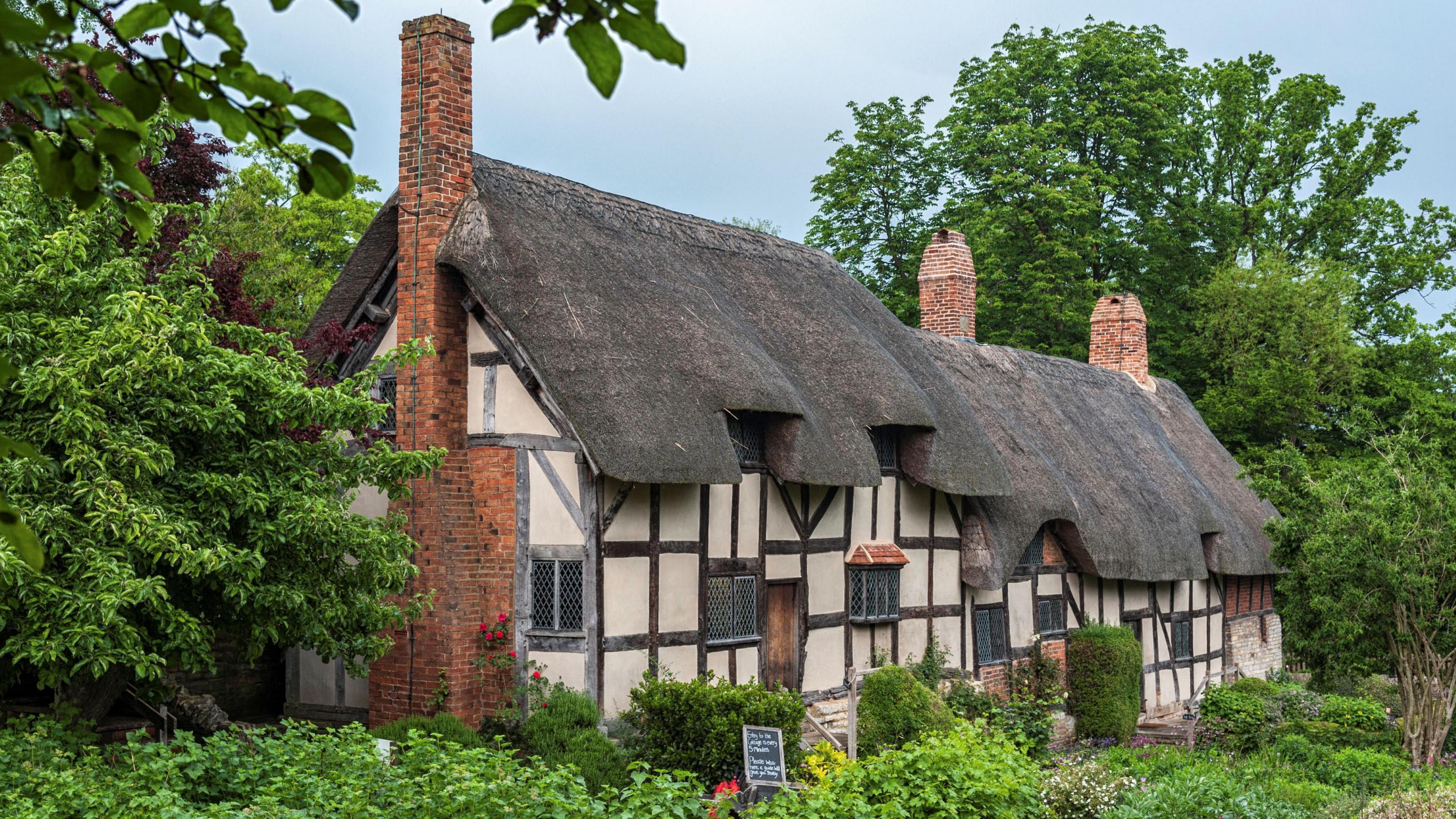 A thatched roof property, with red brick chimneys, leaded windows, timber frame and cream walls. It is surrounded by bushes and trees.