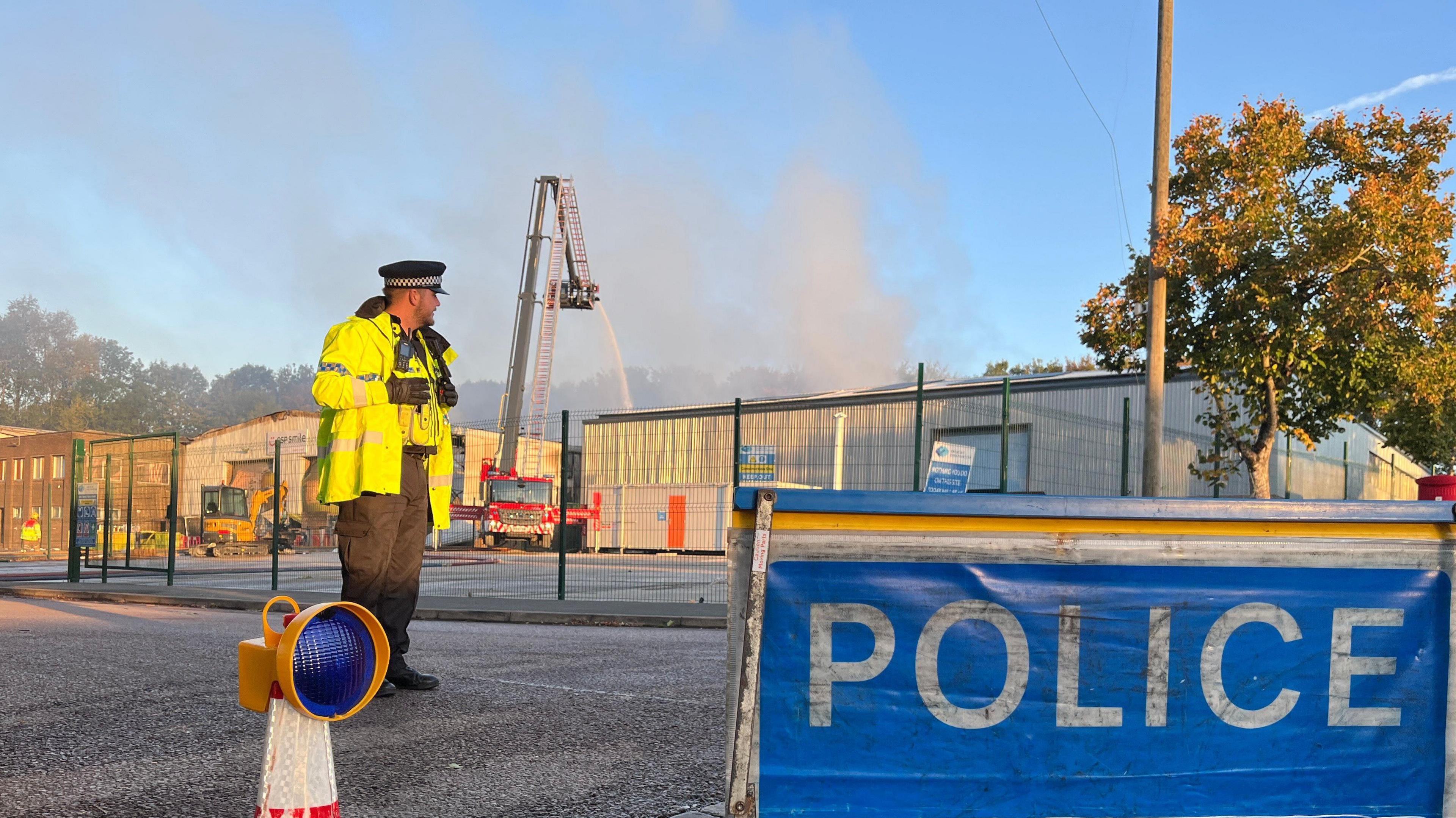 A police officer standing in front of a warehouse at an industrial estate following an explosion and fire. The officer is wearing a black police hat and a high vis jacket. There is a blue police sign on the ground. Smoke can be seen in the background as well as equipment from the fire crews.