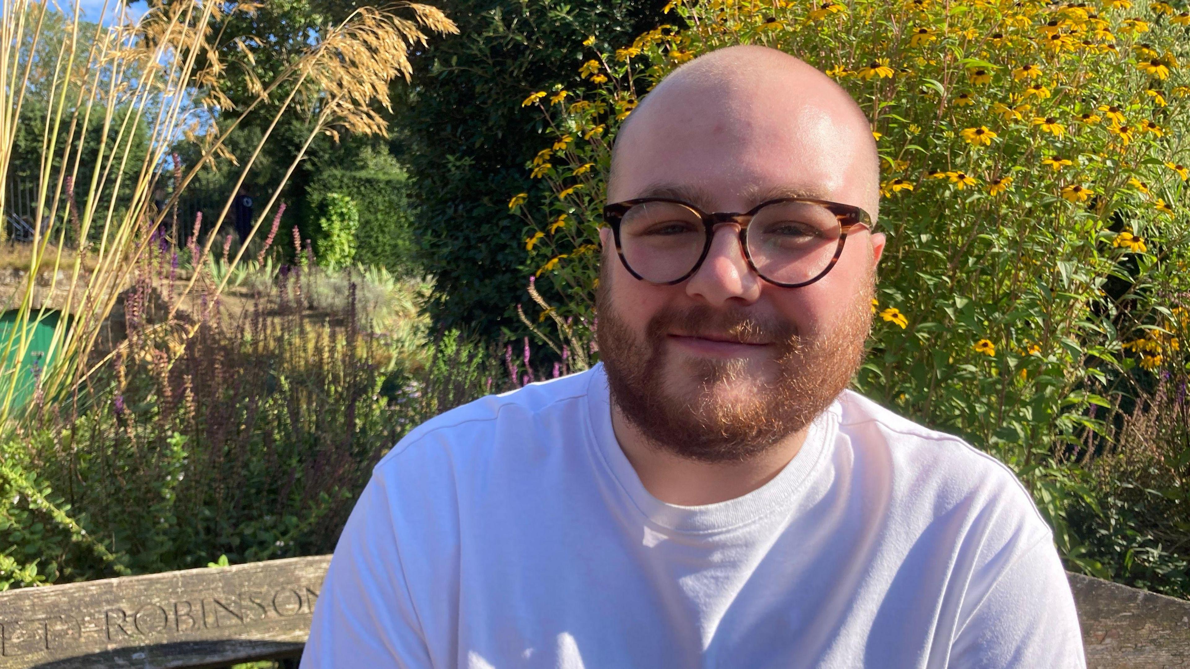 White man with beard and glasses sits on a bench in a garden and looks at the camera. 