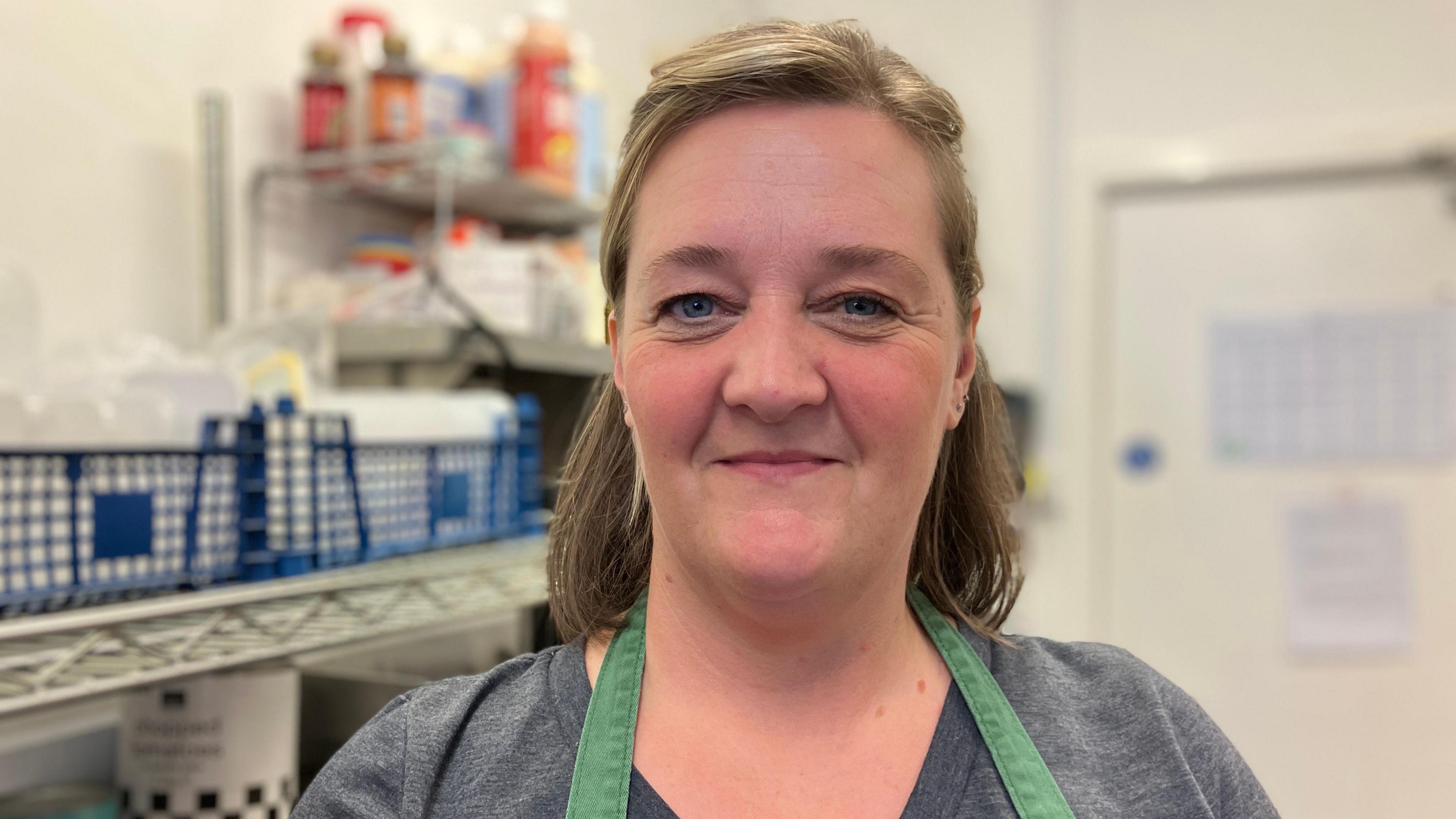 Stacey Bishop wearing a grey t-shirt and a green apron. She has shoulder length light brown hair and is smiling at the camera, standing beside some shelving in a commercial kitchen