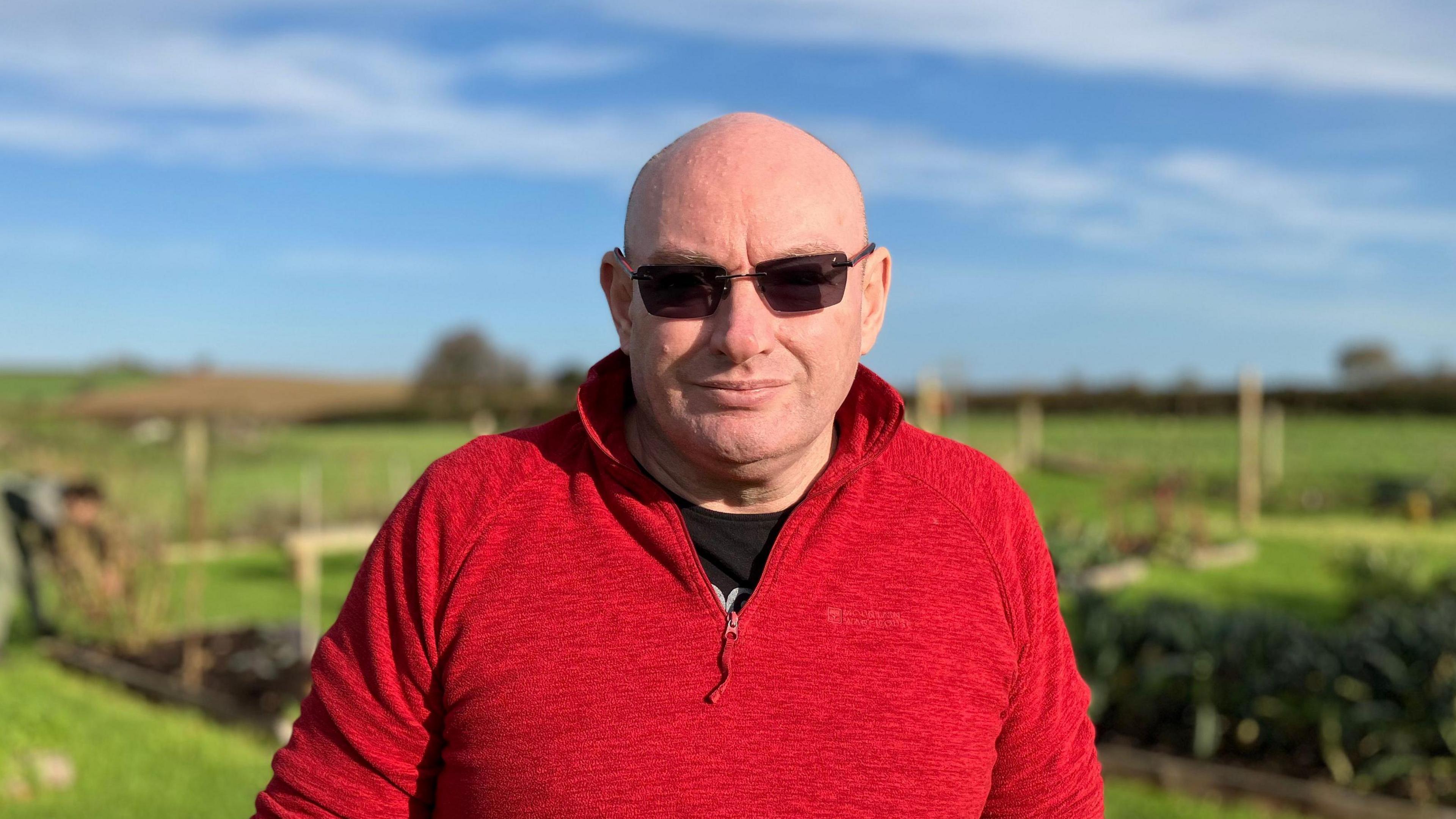 Michael Robins - a bald man in a red fleece with black sunglasses on. He's looking at the camera and has the garden in the background.