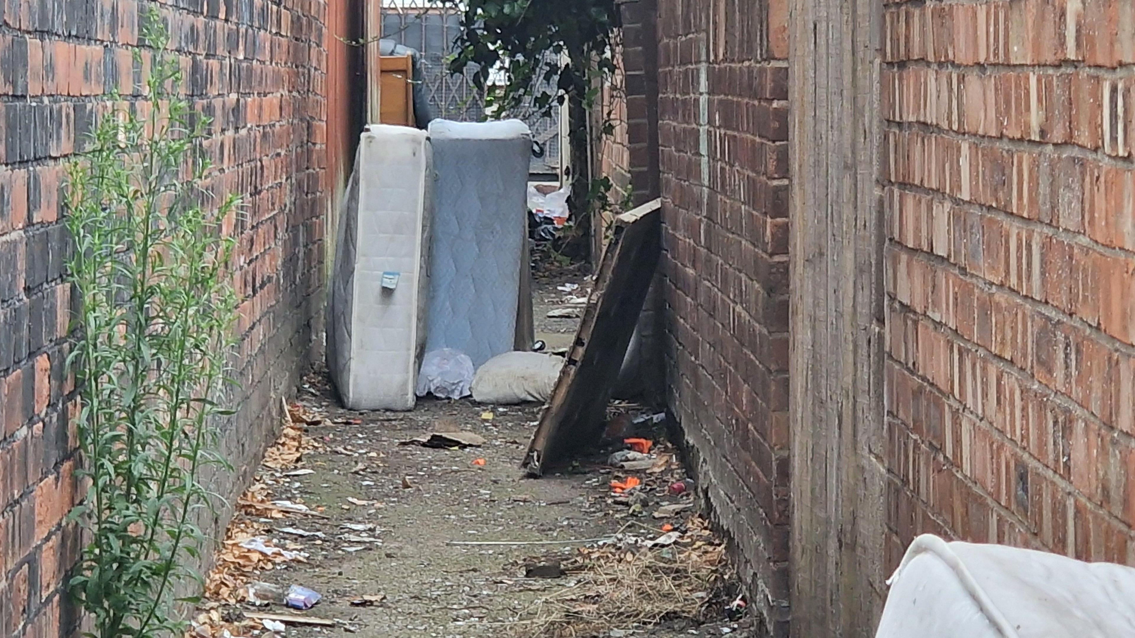 A discarded bed and mattress along with other litter left in a alleyway. There is a third mattress partially in view, a piece of wood and plastic bags.