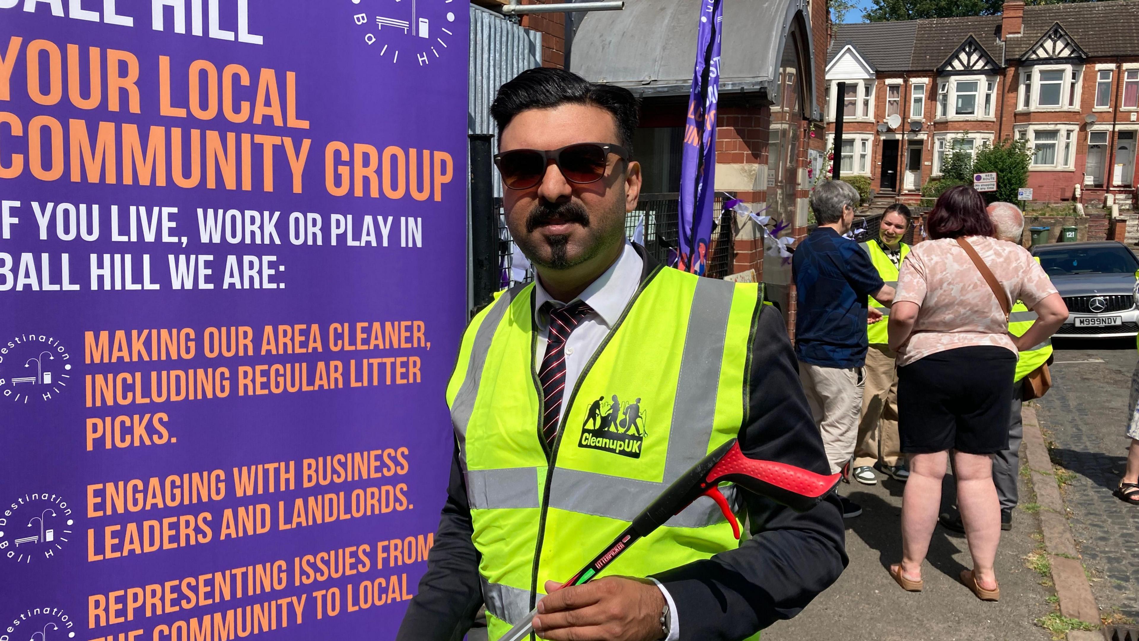 A man in a suit and sunglasses, with a hghi-vis vest, holds a litter picker while standing in front of a purple banner which has the words "YOUR LOCAL COMMUNITY GROUP" in large orange letters.