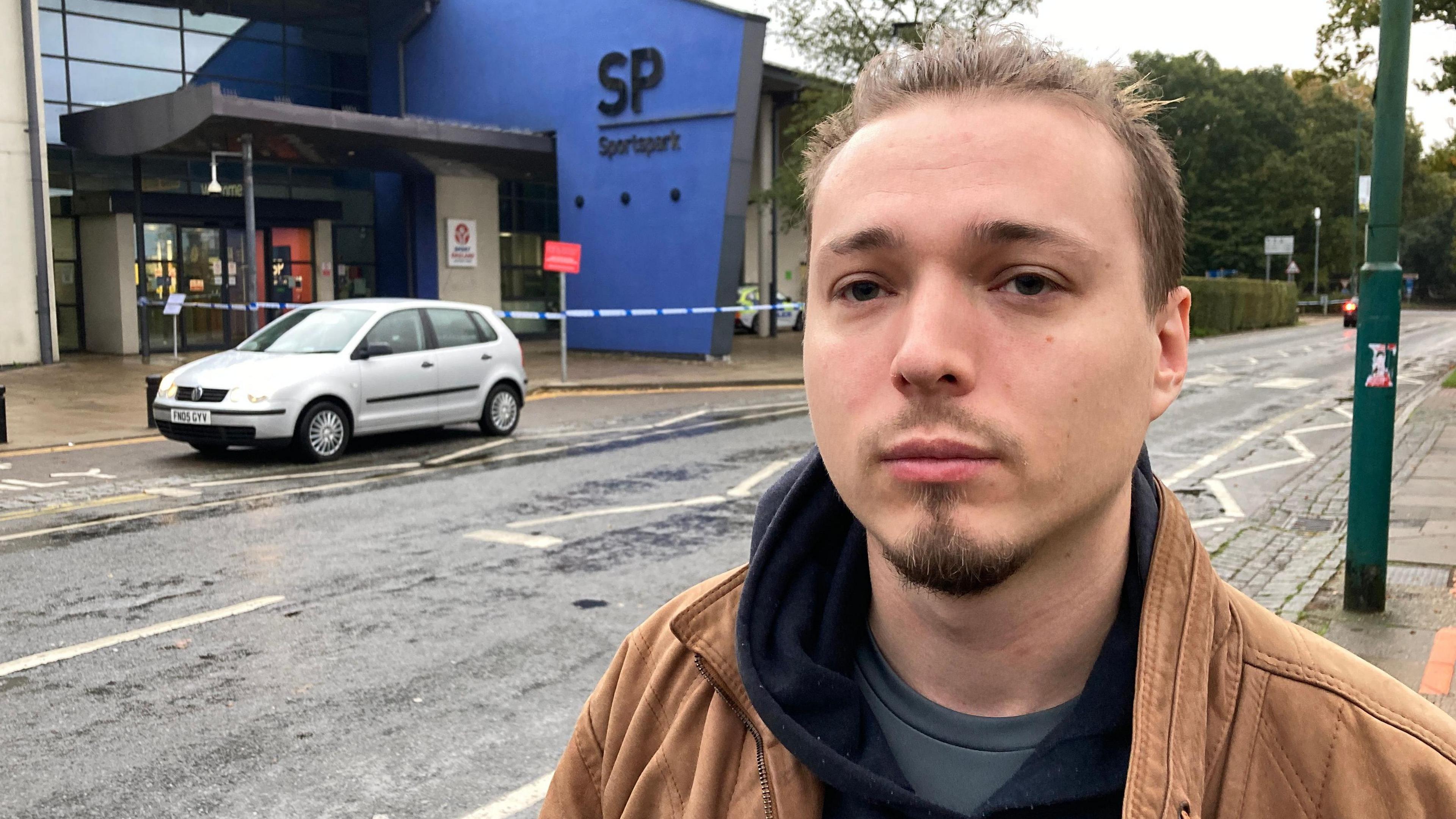 Harry Darlington, who is wearing a brown jacket, black hoodie and grey T-shirt. He has short brown hair and a goatee. He is looking at the camera with a serious expression. In the background is police tape blocking off the entrance to the Sportspark, which is a blue and white building.