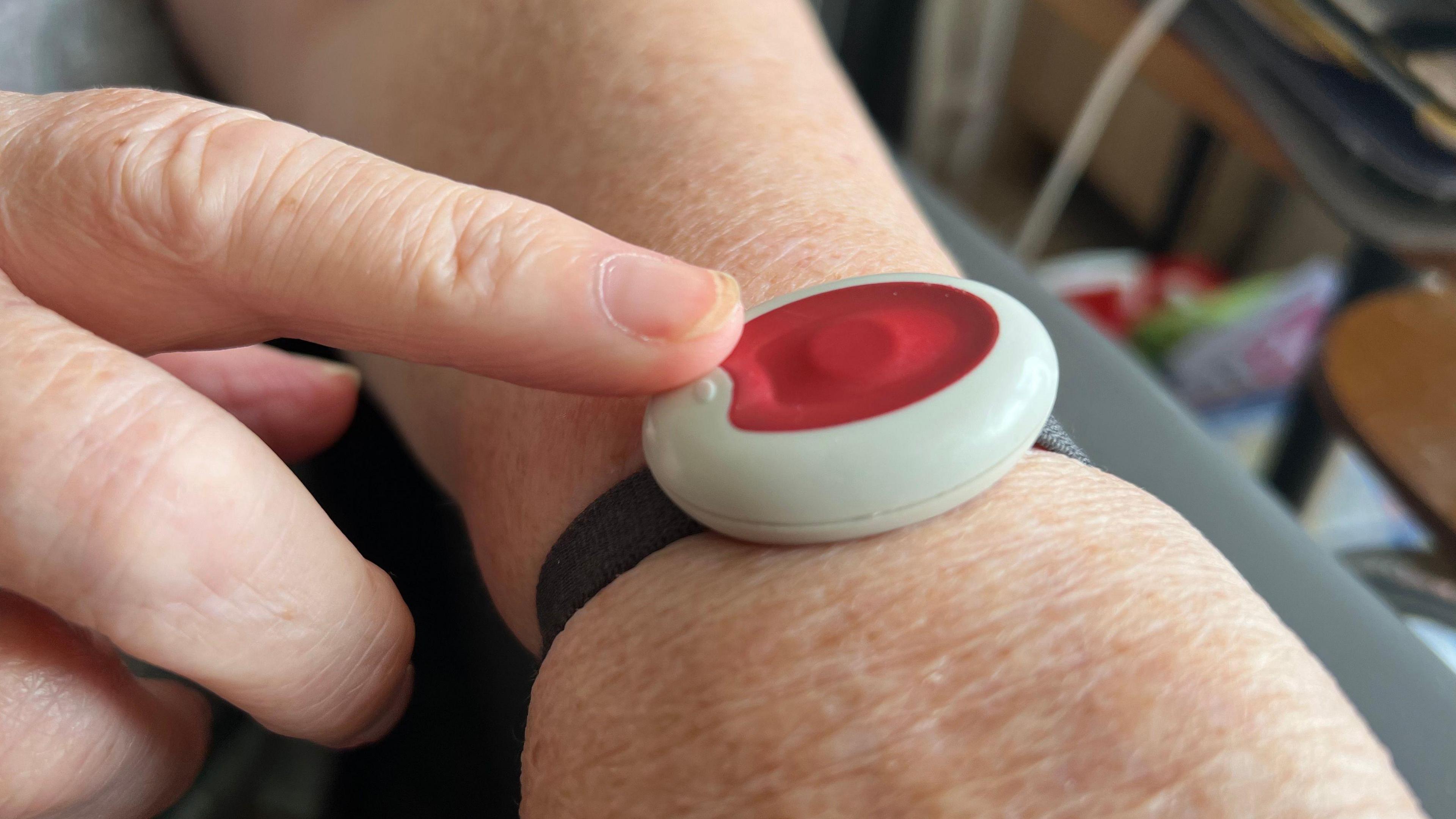 Photo of a red alarm button strapped to a wrist with a black strap with a finger touching it