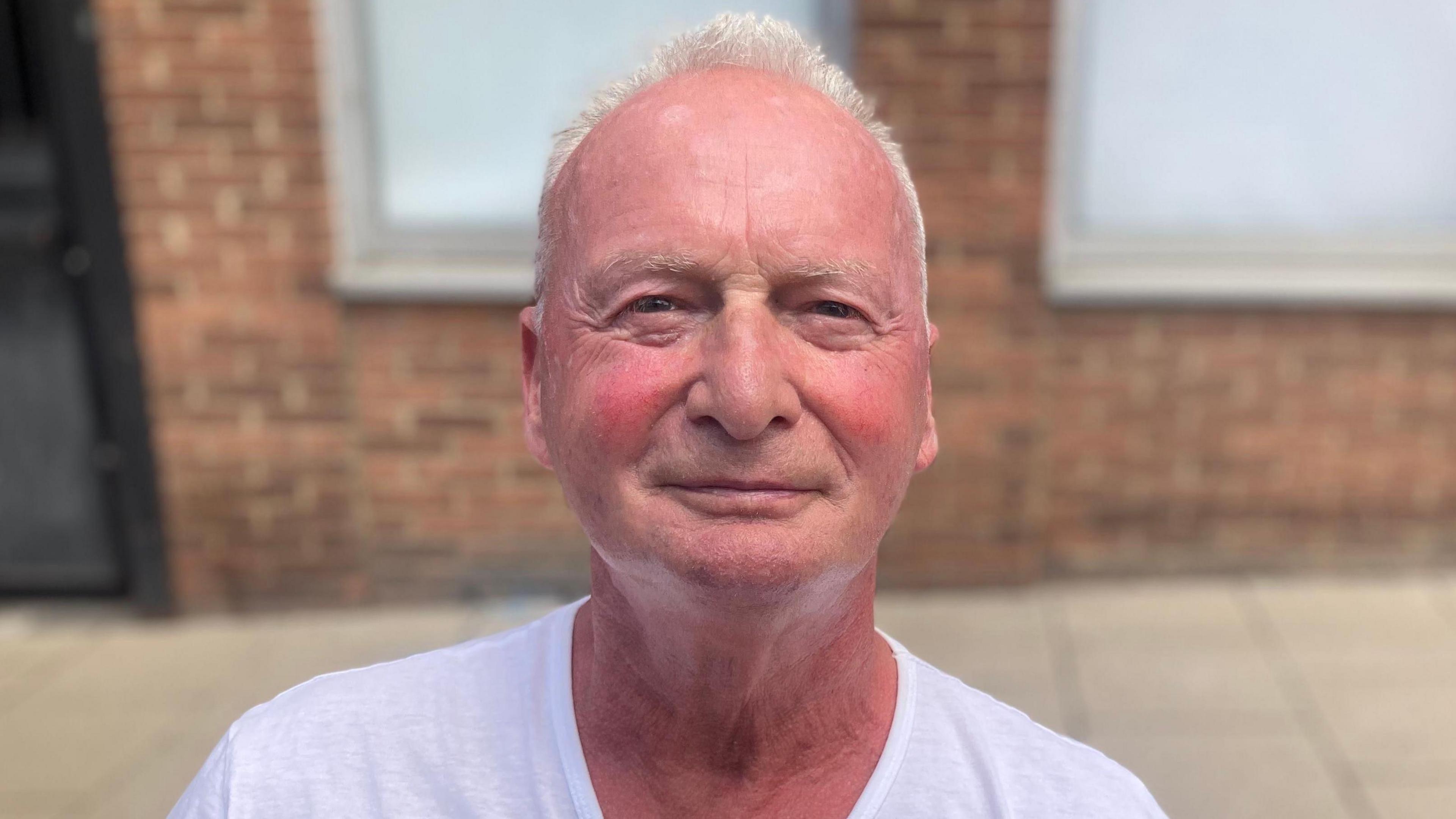 Paul Davis is looking at the camera with a light smile. He is wearing a white T-shirt and has short grey hair. He is standing in front of a red bricked building.