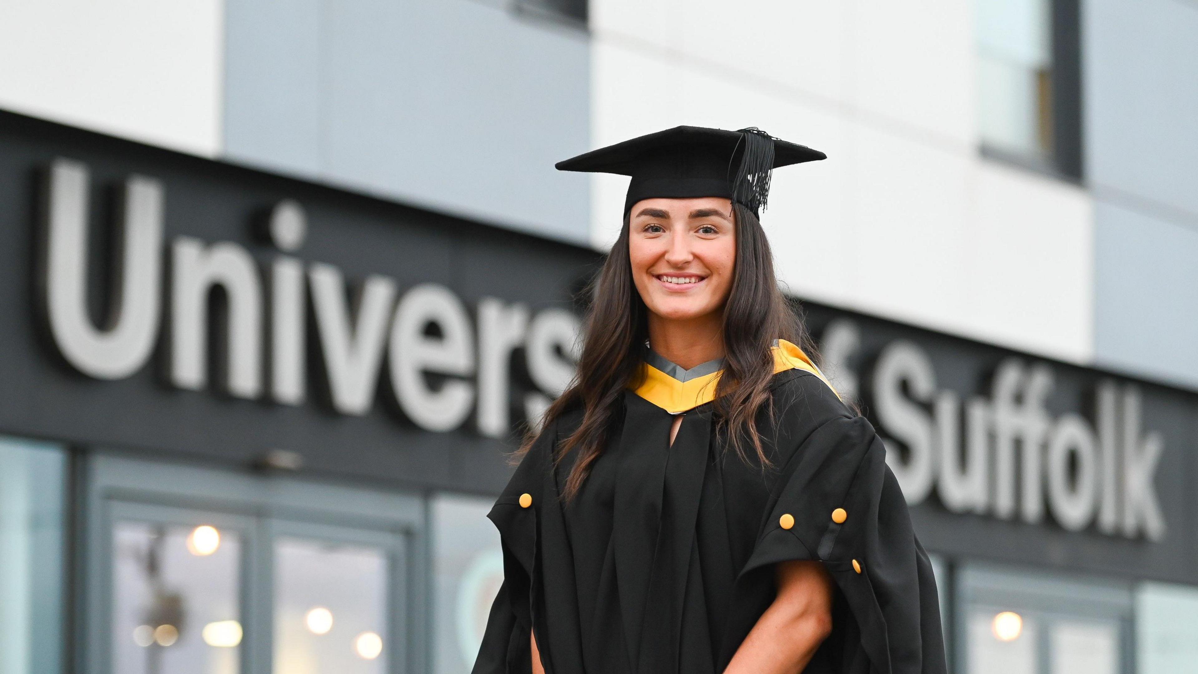 A woman, who is wearing a black and yellow academic robe and a black mortar board. standing in front of a blurred University of Suffolk sign  