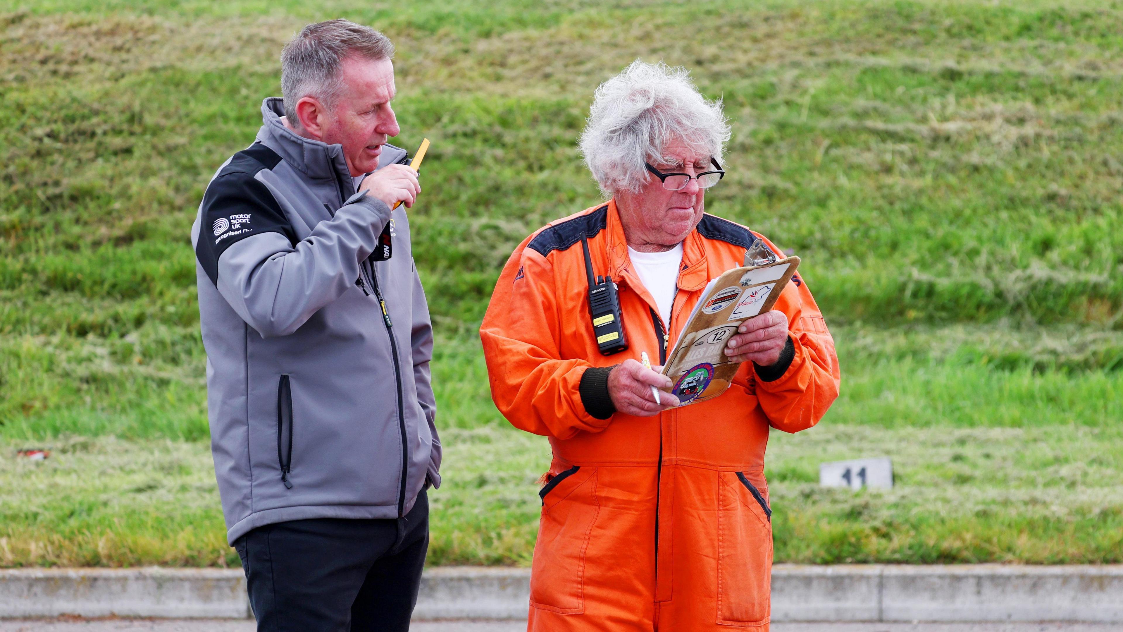 Maurice Bennington, standing to the left wearing an orange jumpsuit next to a man wearing a grey top and dark trousers, talking into a walkie-talkie. Maurice has grey hair and glasses, and is holding a clip-board and pen, the other man is looking at the clip-board. There is grass behind them. 