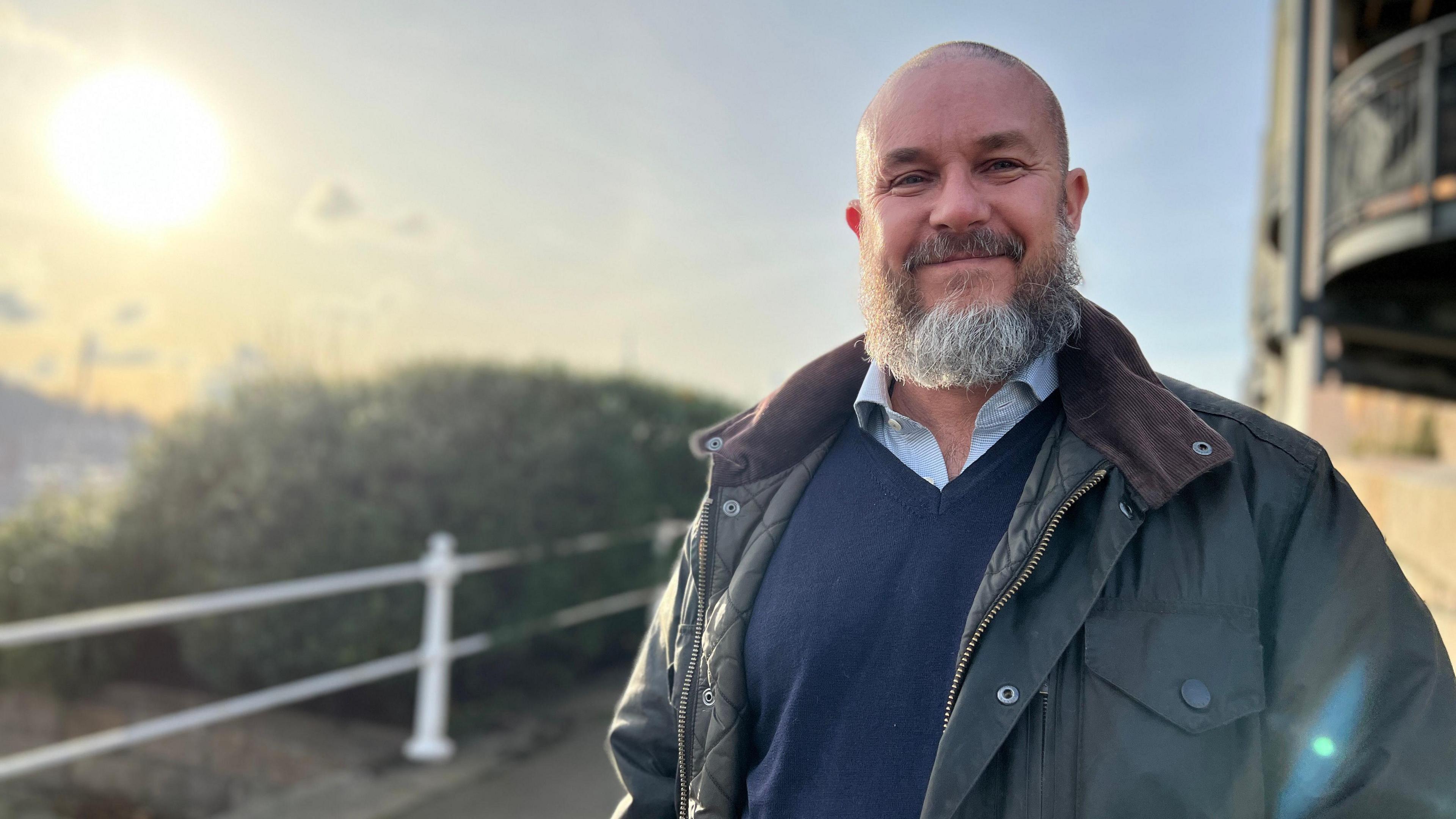 A bald man with a beard smiles. He is wearing a blue shirt and v-necked jumper and a green wax jacket, and stands with the sun behind him behind him next to some railings.