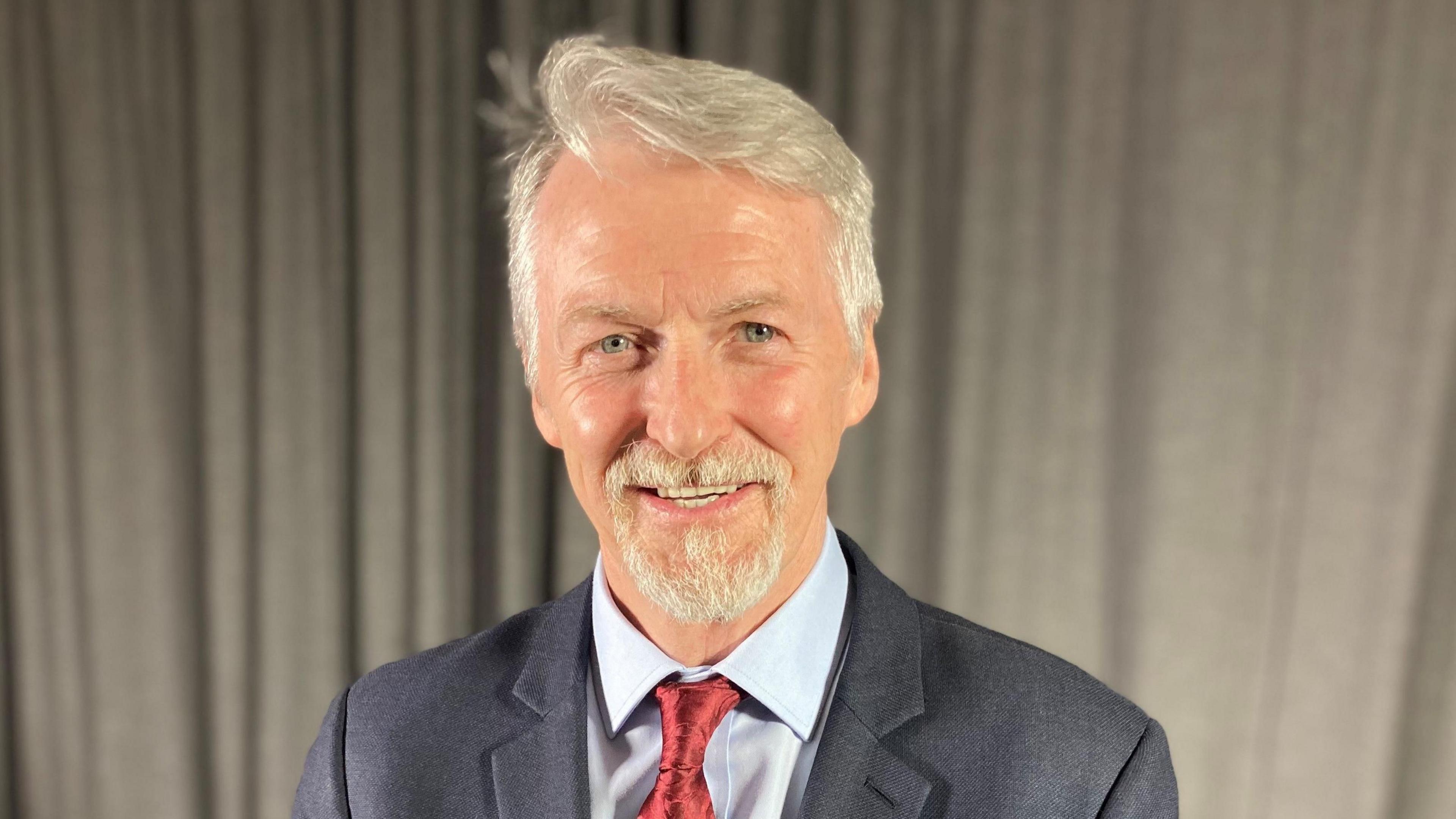 Huw Irranca-Davies, who has grey hair and  a goatee beard smiles at the camera. He is wearing a grey suit and red tie against a pale shirt. Behind him is a beige curtain