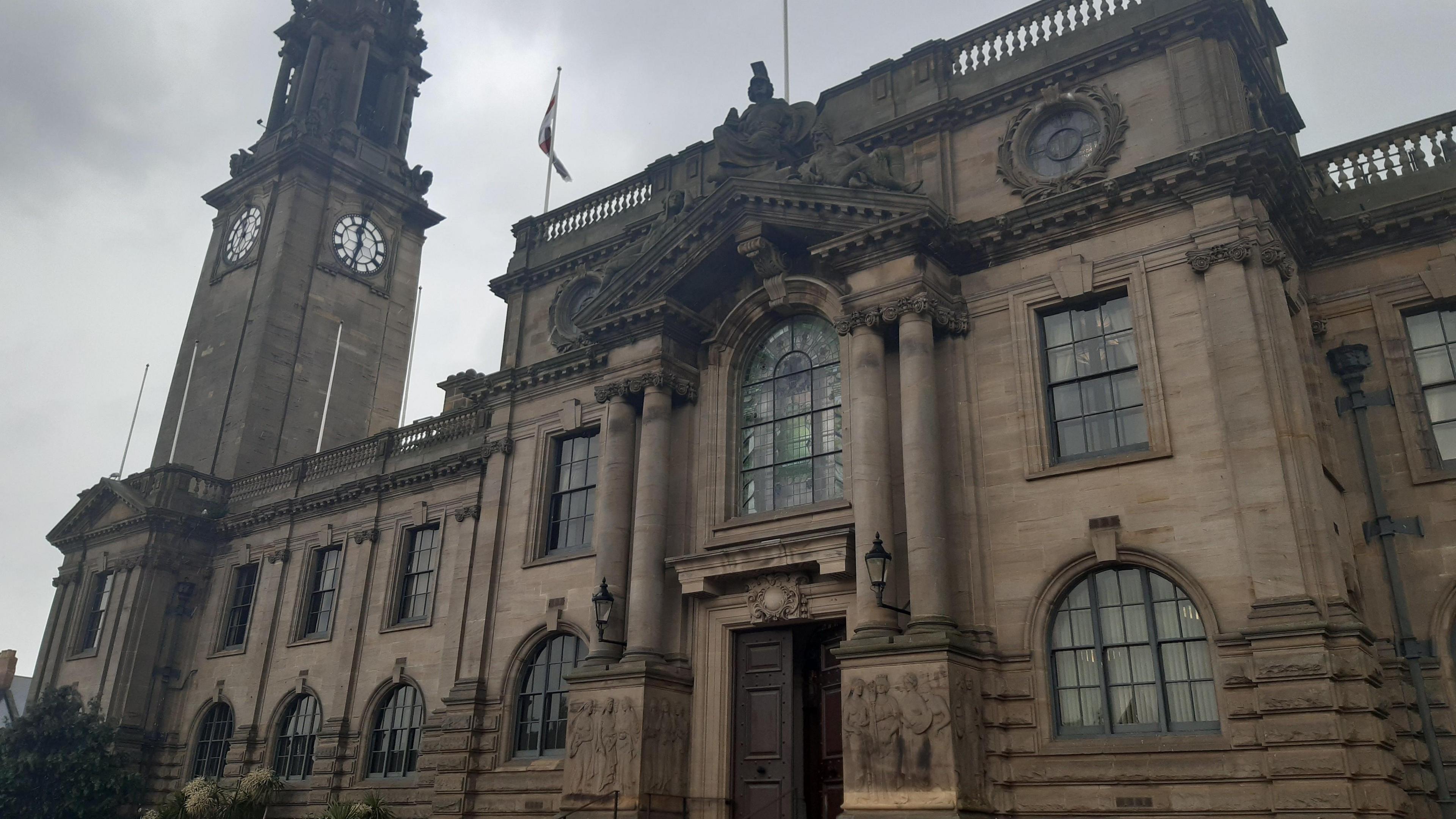 South Shields Town Hall, a large stone building with tall windows, carved pillars, and a sculpted depiction of a knight atop an arched roof. There is a block tower to the left and two flag poles on the roof. The sky above is grey and cloudy.