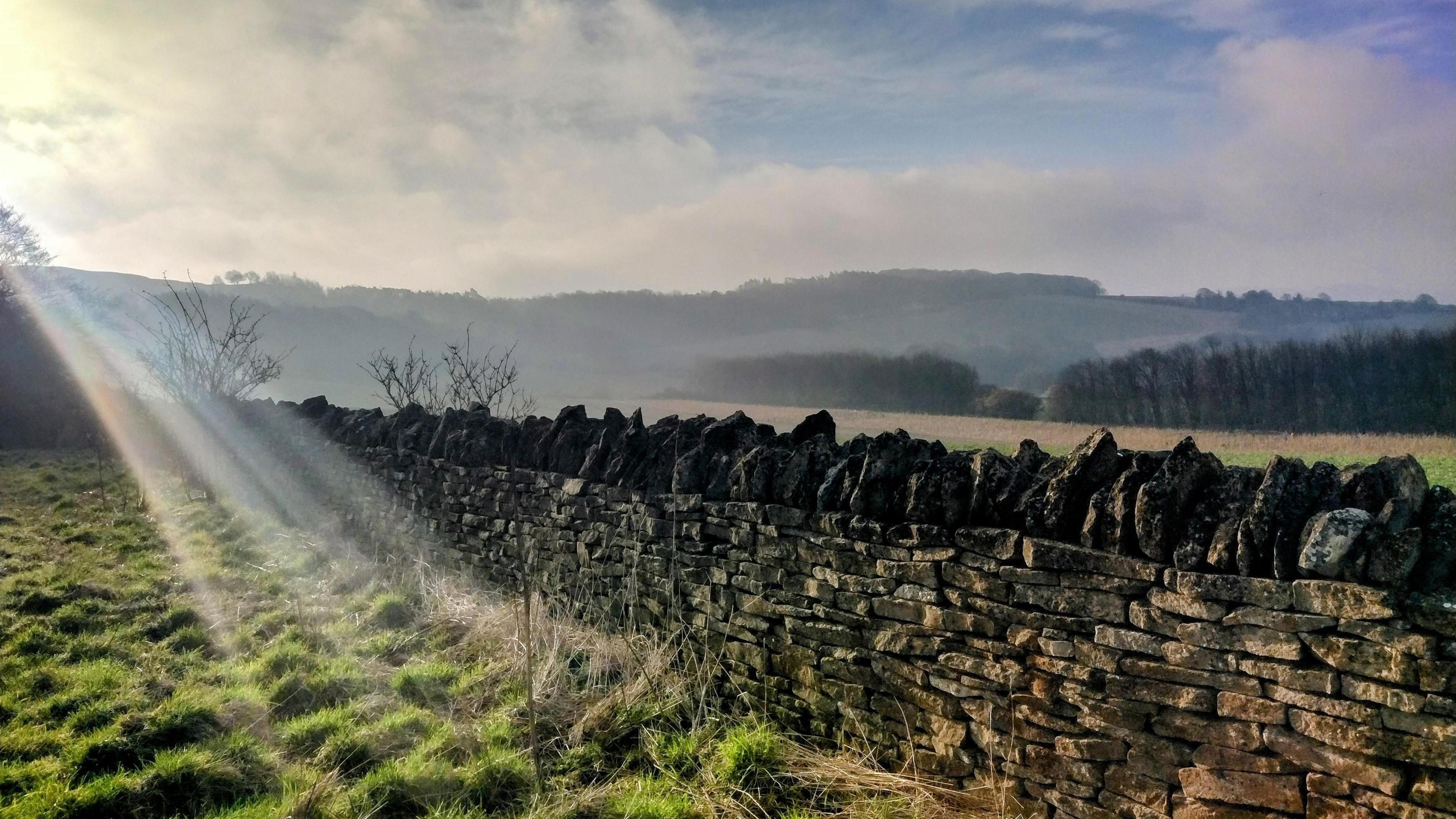 A sun ray creeping into the picture shining on a stone wall in the countryside with a misty background under a blue sky. 