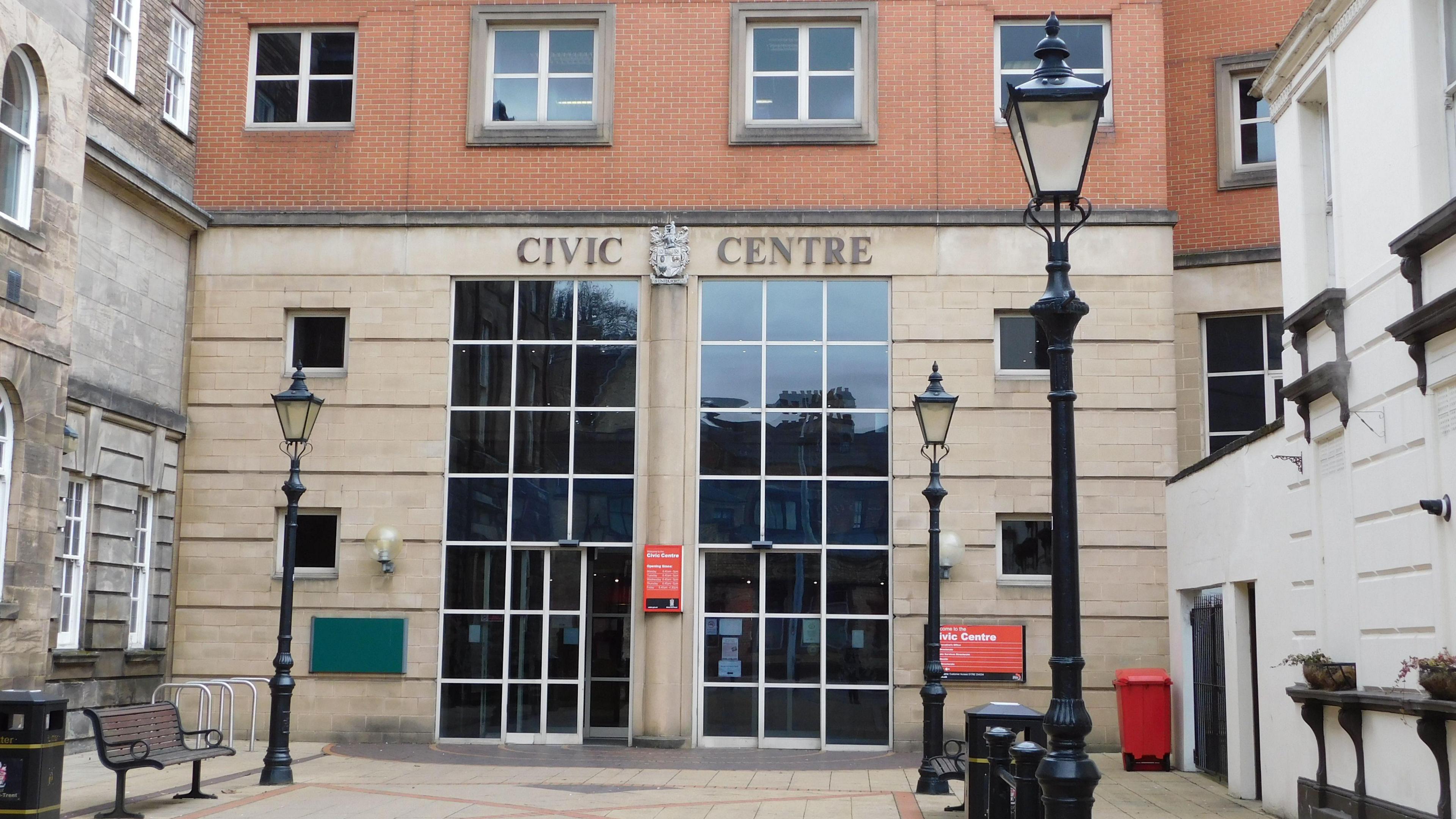 A large brick and beige building with dozens of glass windows on the front and lamp posts and benches outside.